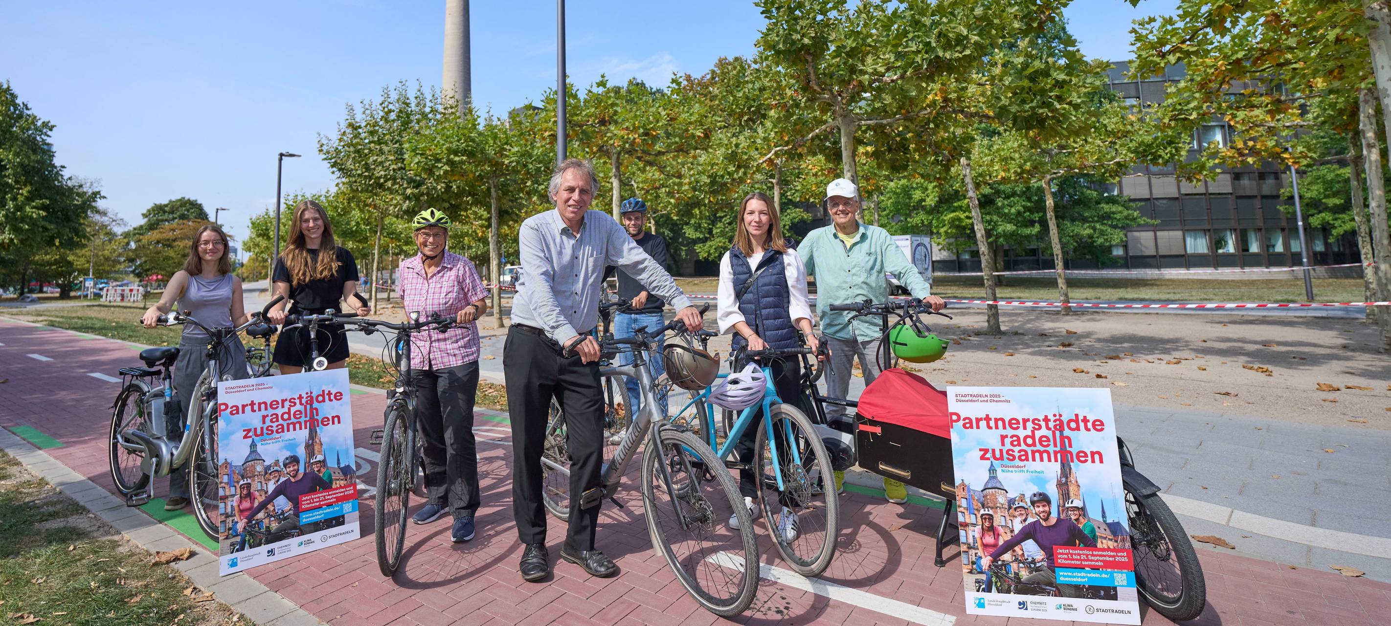 Jochen Kral (M.), Dezernent für Mobilität und Umwelt, und Mitarbeiter der Stadt präsentieren das Stadtradeln-Plakat. Sie stehen neben ihren Fahrrädern. Im Hintergrund ist der Rheinturm zu sehen.