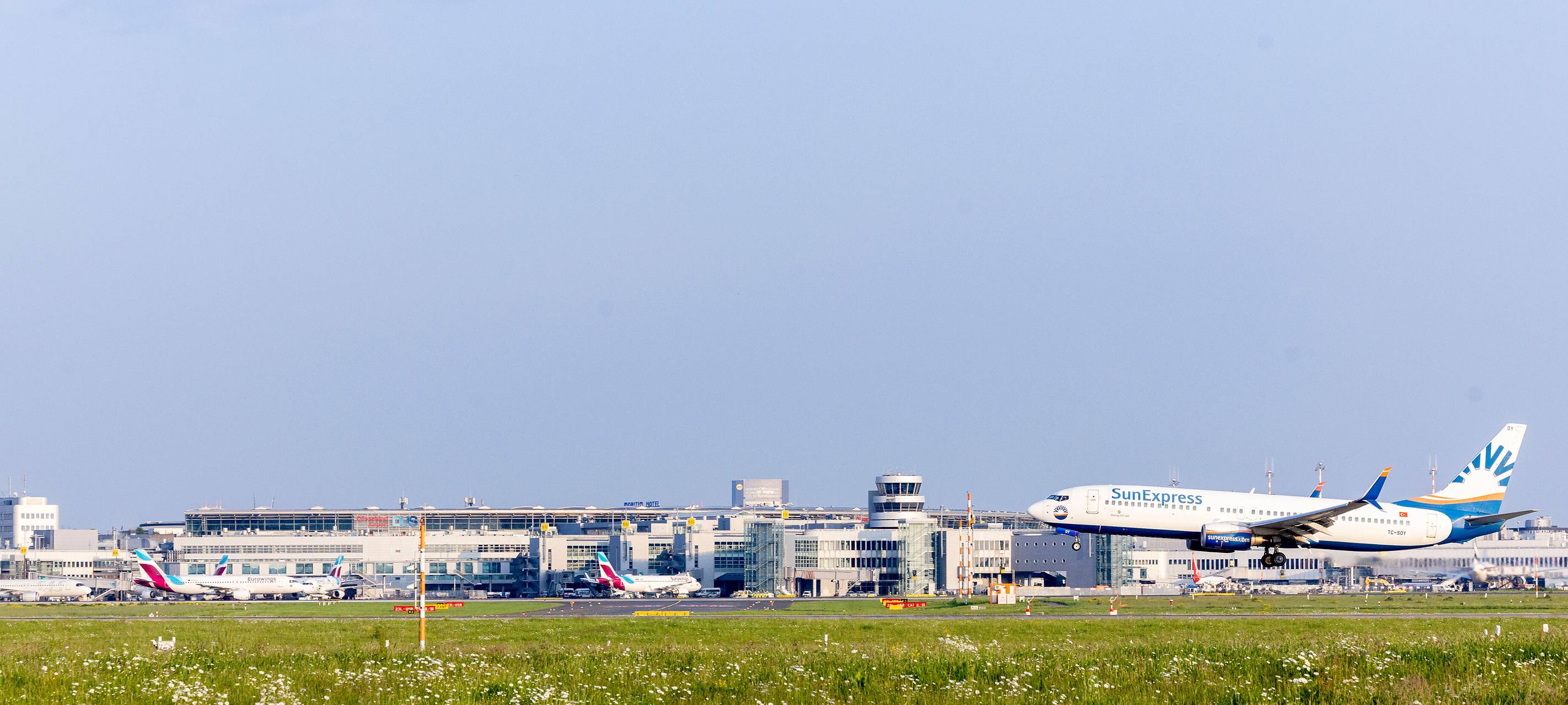 Rechts im Bild ein Flugzeug auf der Startbahn, das gerade abhebt, davor Rasen. Im Hintergrund der Düsseldorfer Flughafen, davor stehen einige Flugzeuge.