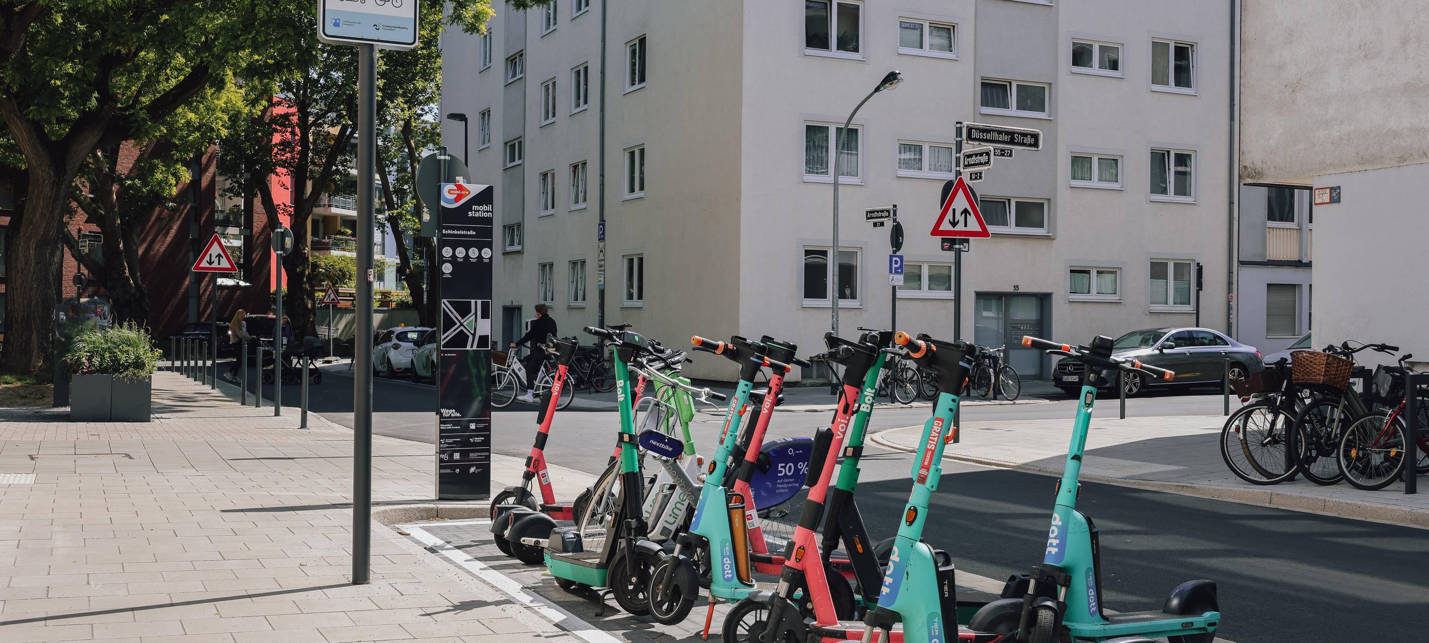 Ein paar Leihroller stehen an einer Mobilitätsstation in Düsseldorf. Im Hintergrund sind eine Straße und Hochhäuser