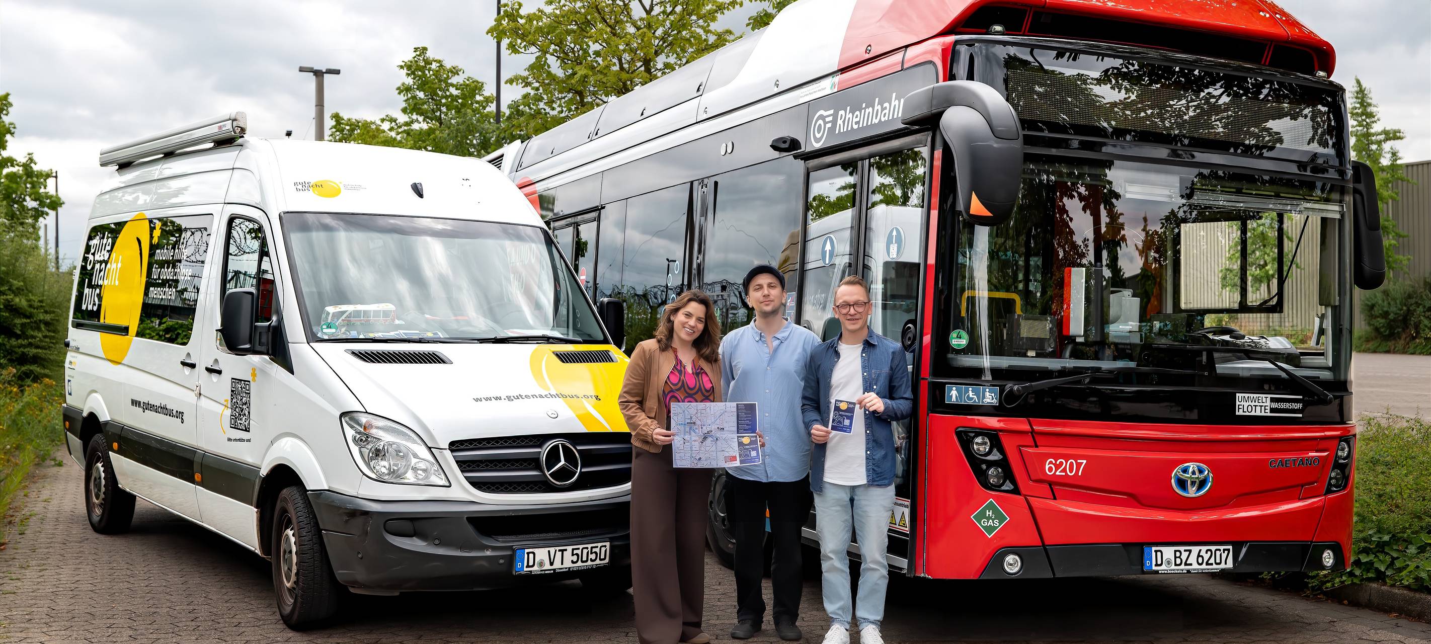 Vor einem weißen Kleinbus und einem roten Linienbus stehen drei Menschen und lächeln.