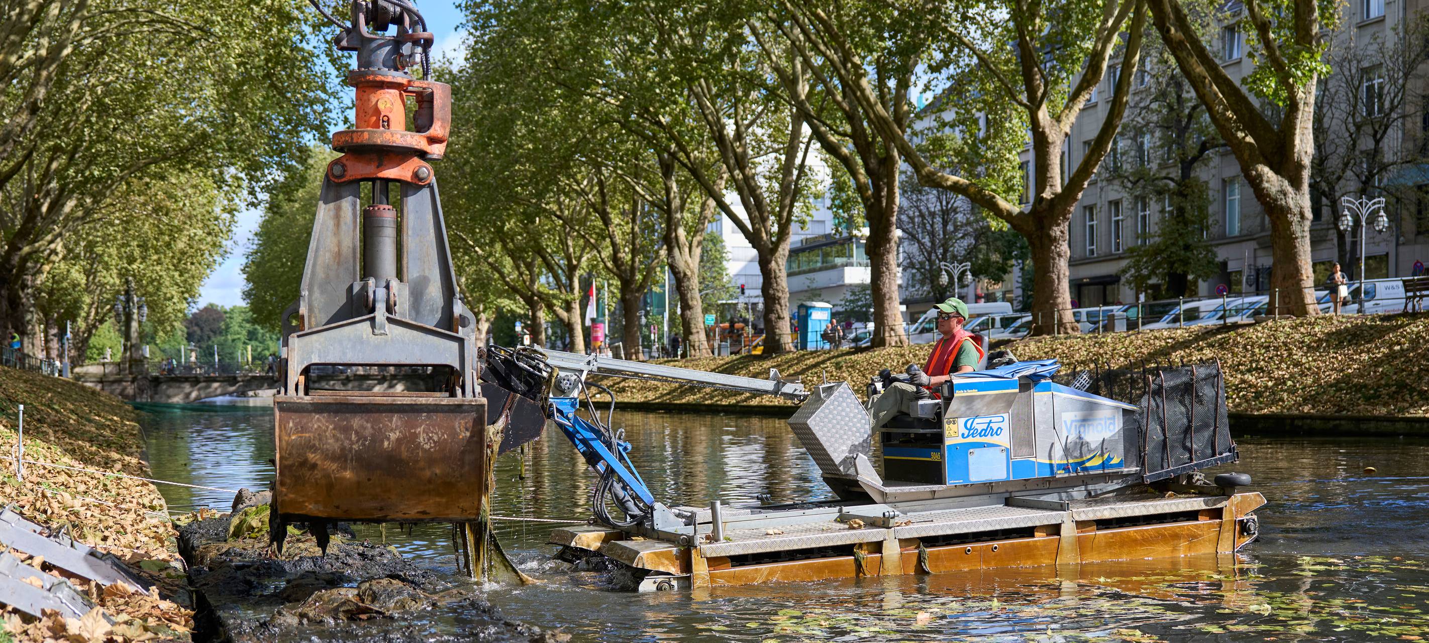 Ein Amphibienfahrzeug steht im Wasser eines Grabens und schiebt Schlamm an den Rand. Ein Bagger hebt den Schlamm heraus.