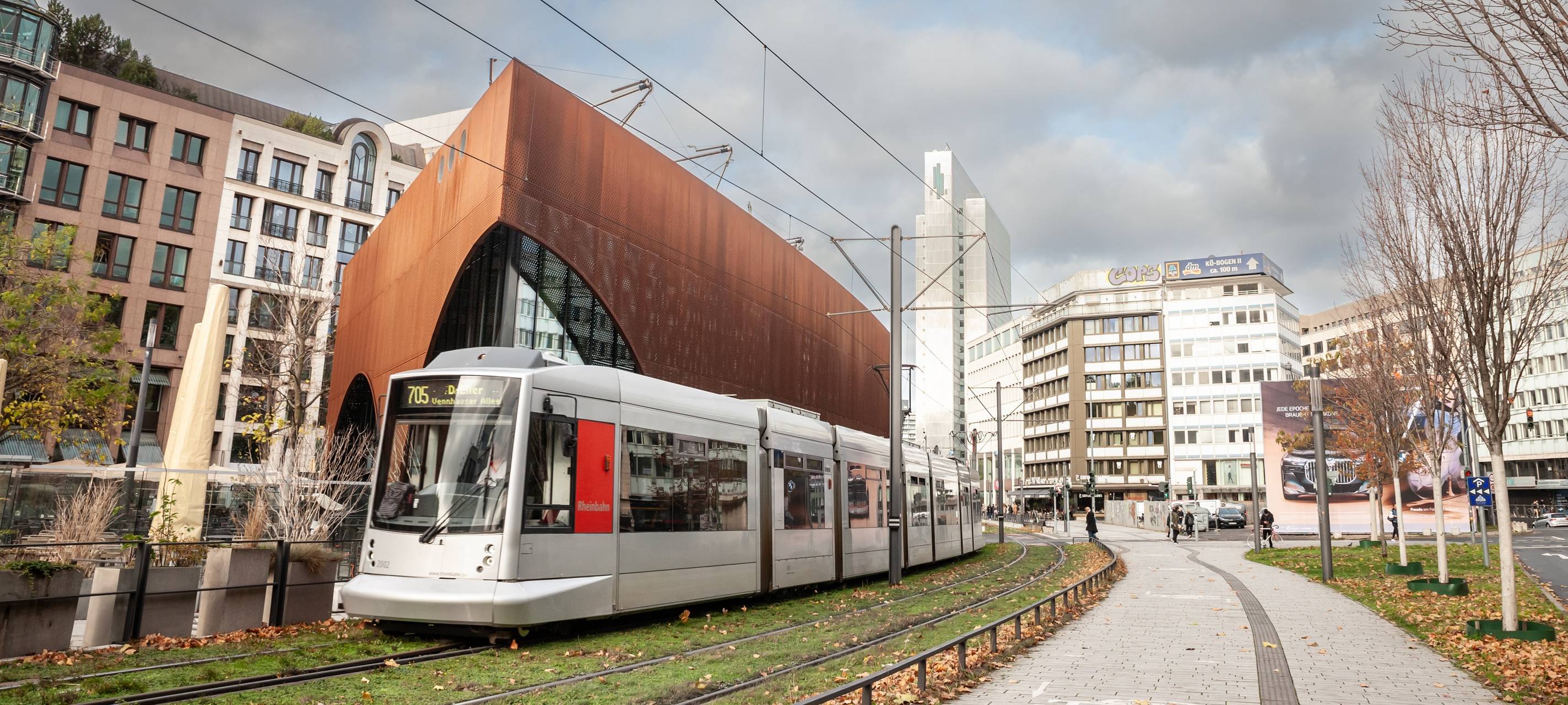 Eine Straßenbahn der Düsseldorfer Straßenbahn fährt durch das Stadtzentrum von Düsseldorf. Die Bahn wird von der Rheinbahn betrieben und ist Teil des öffentlichen Nahverkehrs. Im Hintergrund sind städtische Gebäude und Passanten zu sehen.