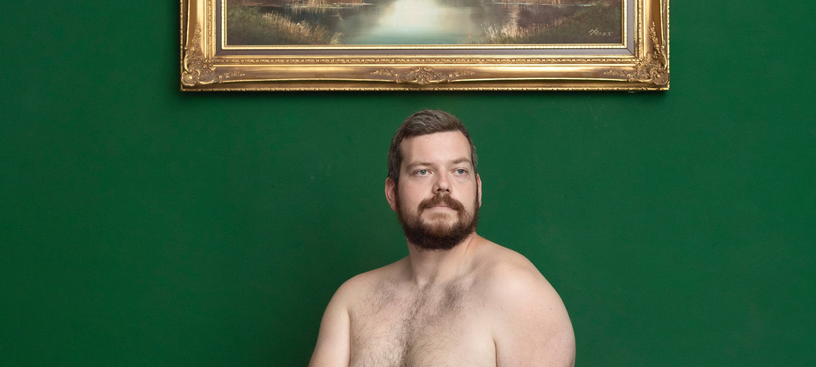 Ein nacker Mann sitzt vor einem Bild. Dahinter eine grüne Wand.