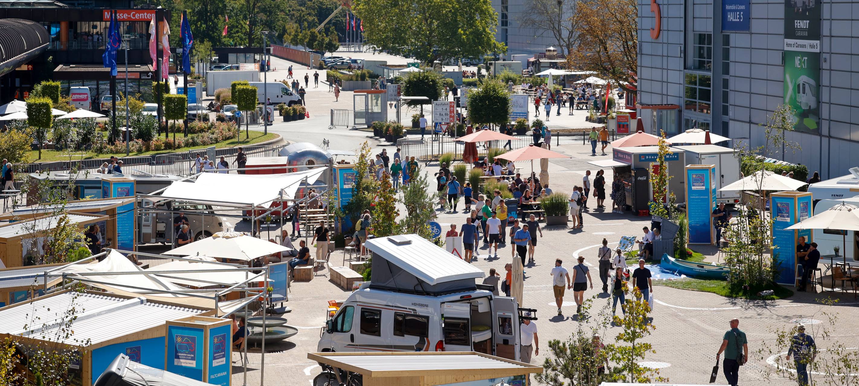 Ein Parkplatz mit Gastro-Zelten und Caravans. Zwischen den Ständen laufen Menschen hin und her.