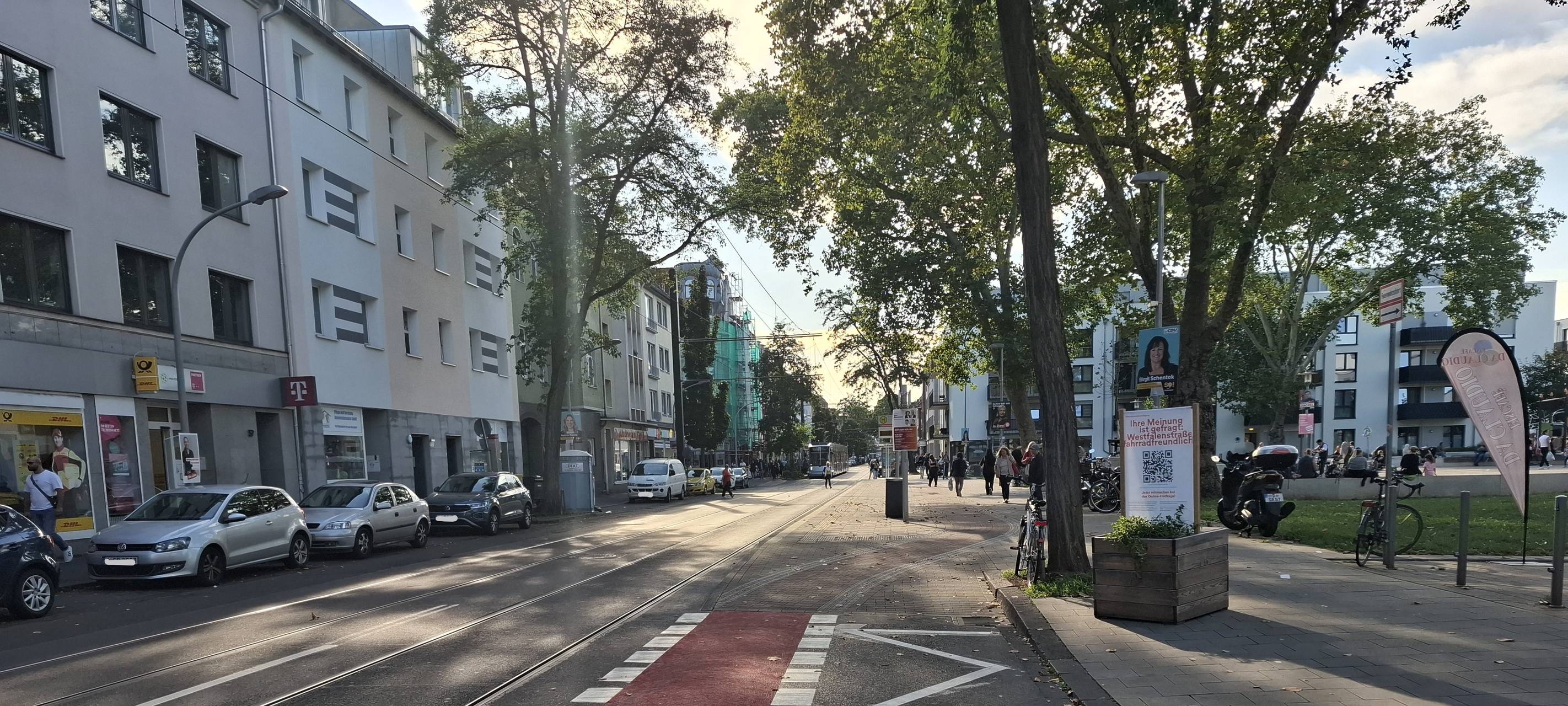 Ein roter Fahrradstreifen am rechten Straßenrand, rechts stehen Bäume, links sind parkende Autos und Häuser zu sehen.