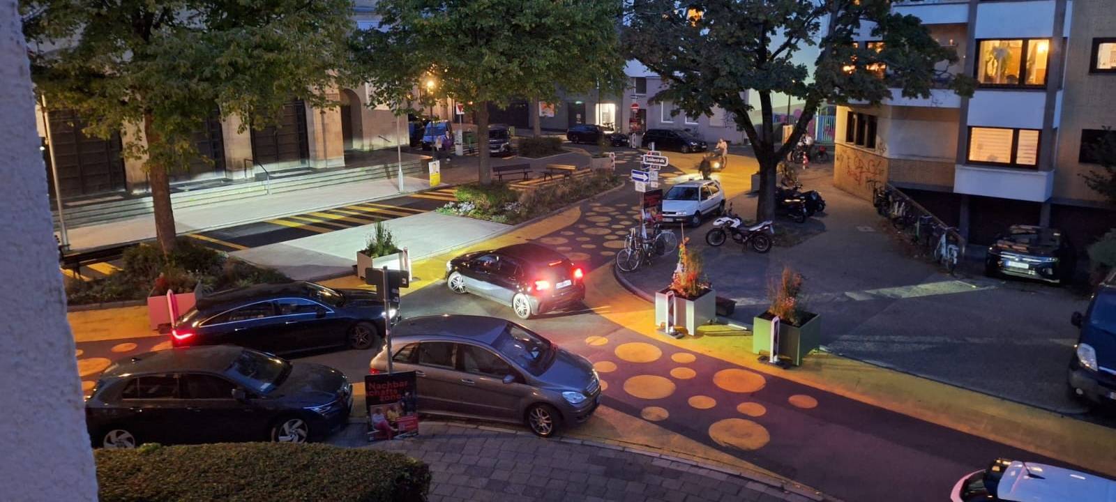 Auf dem Bild sieht man den Paulusplatz mit gelben Streifen und Punkten auf der Straße. Zusätzlich hat die Stadt Blumenkübel und Warnbaken aufgestellt. Mehrere Autos versuchen an den Kübeln vorbeizufahren.