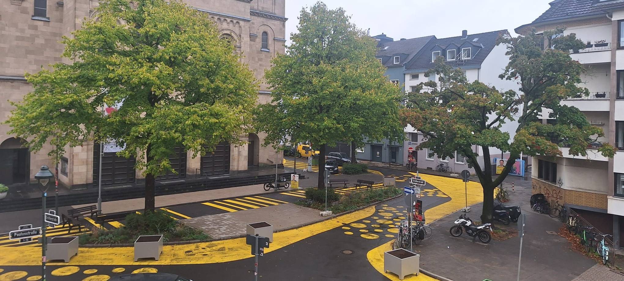 Auf dem Bild sieht man den Paulusplatz mit gelben Streifen und Punkten auf der Straße. Zusätzlich hat die Stadt Blumenkübel aufgestellt.