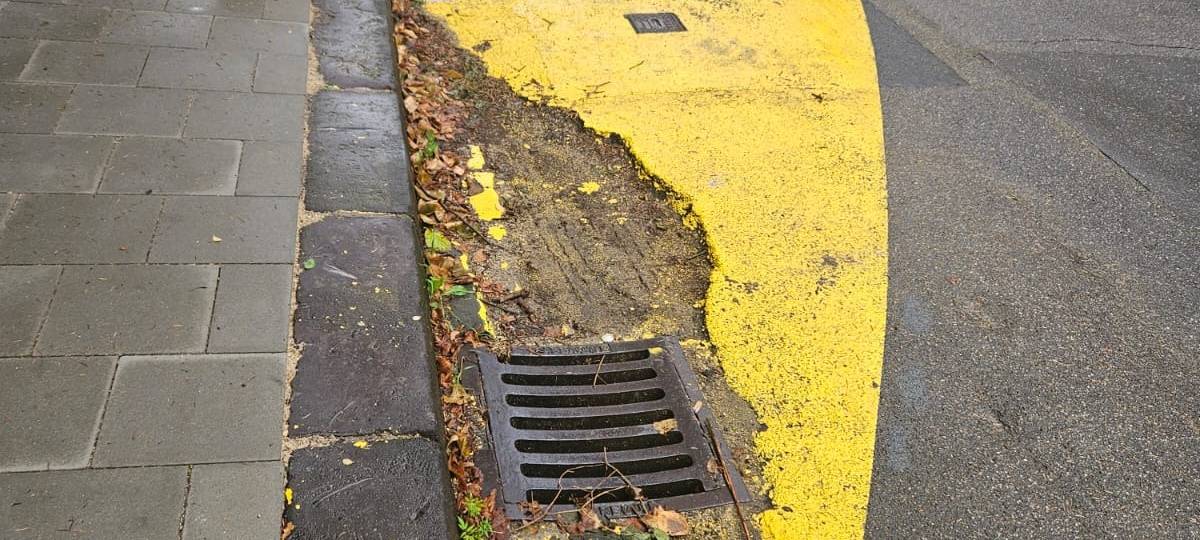 Auf dem Bild ist ein Stück Straße am Paulusplatz zu sehen. Es ist mit gelber Farbe bemalt. Die Farbe und der Straßenbelag bröckeln.