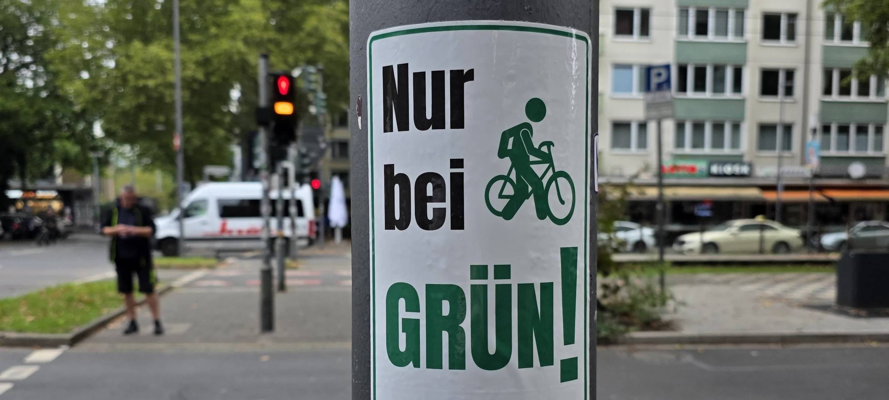 Ein grün-weißer Aufkleber mit dem Slogan "Nur bei Grün! – Vorbild sein!" klebt auf einem Ampelmast. Im Hintergrund ist eine Kreuzung zu sehen.