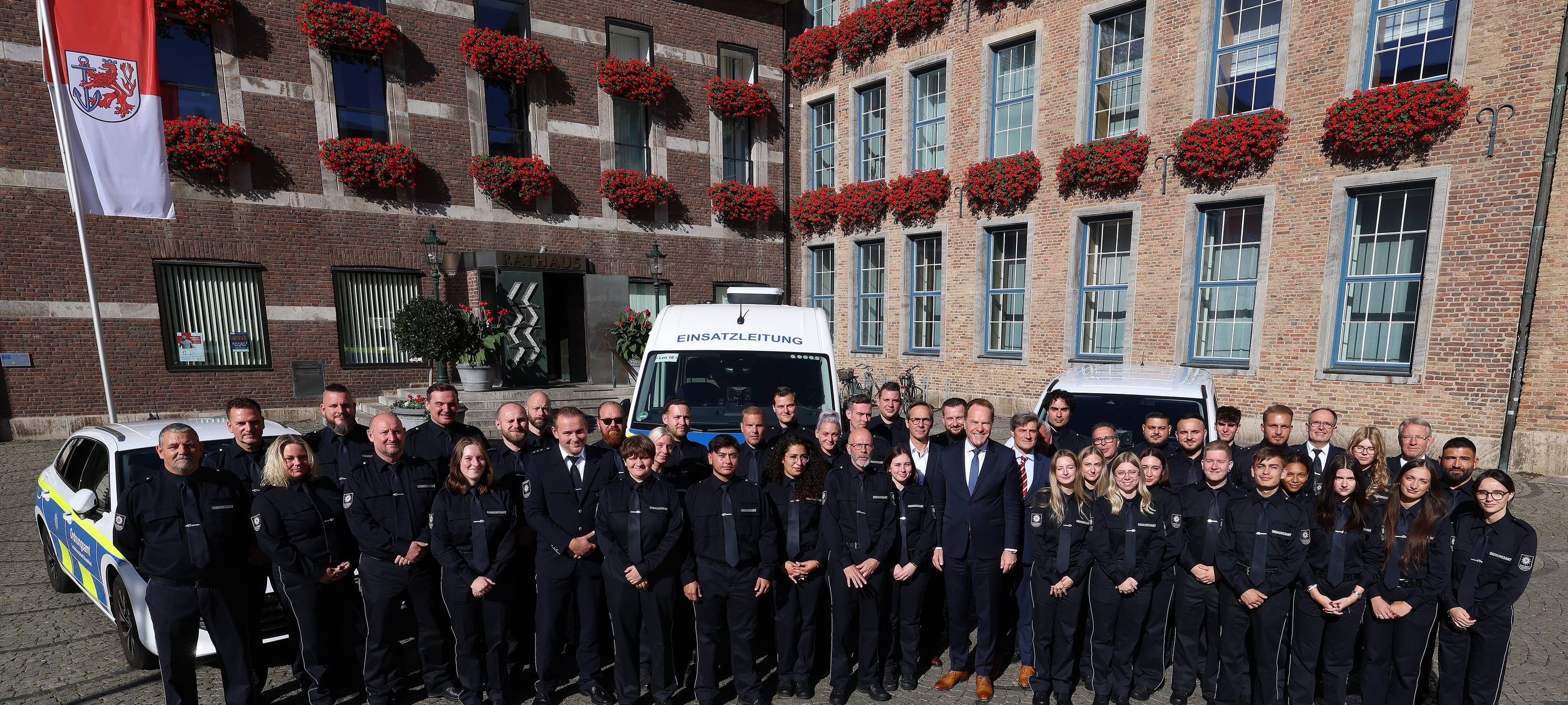 Über 50 neue OSD-Mitarbeitende stehen aufgereiht vor dem Düsseldorfer Rathaus. Hinter der Gruppe parken Autos des OSD. Vor dem Rathaus weht die Düsseldorf-Flagge.