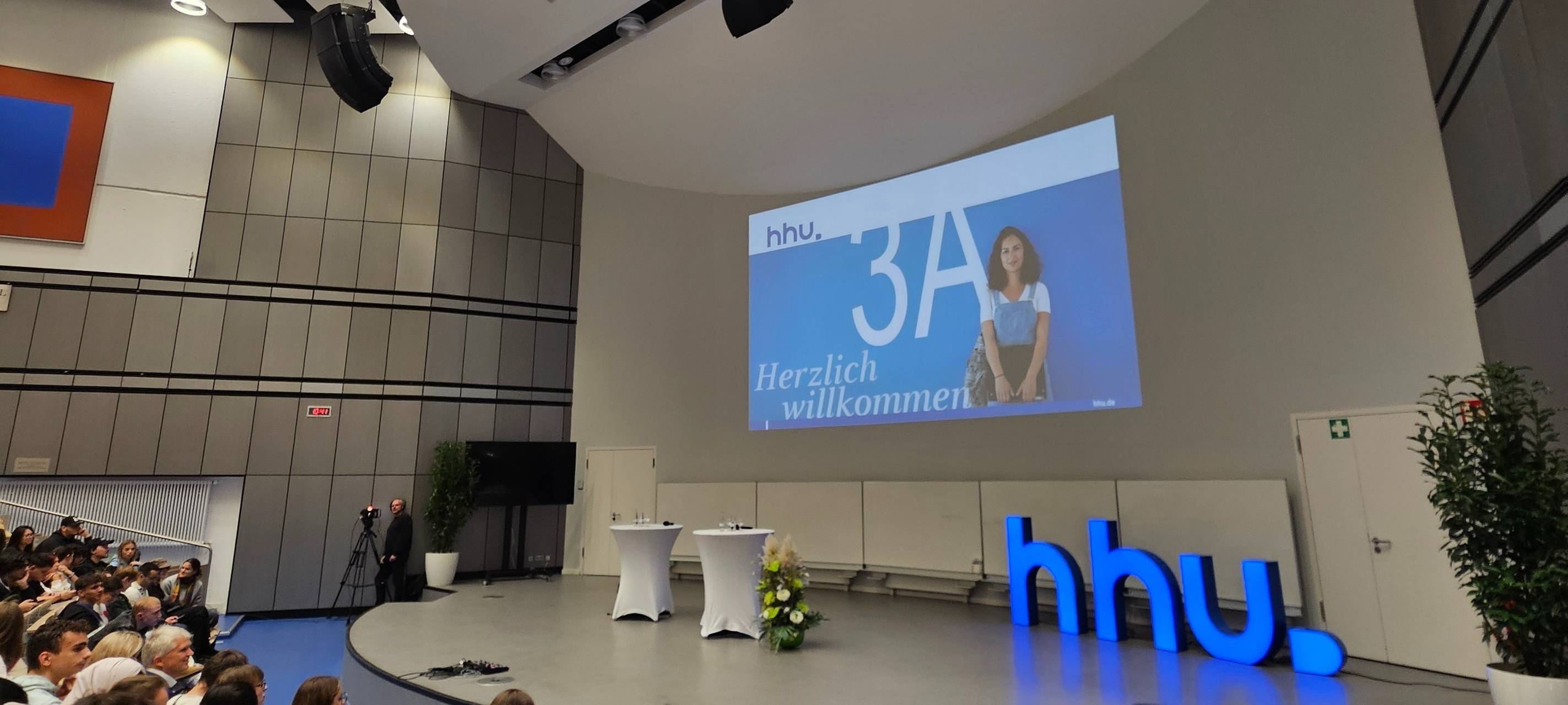 Bühne im Hörsaal 3A der Heinrich-Heine-Universität mit hhu Zeichen auf der rechten Seite.