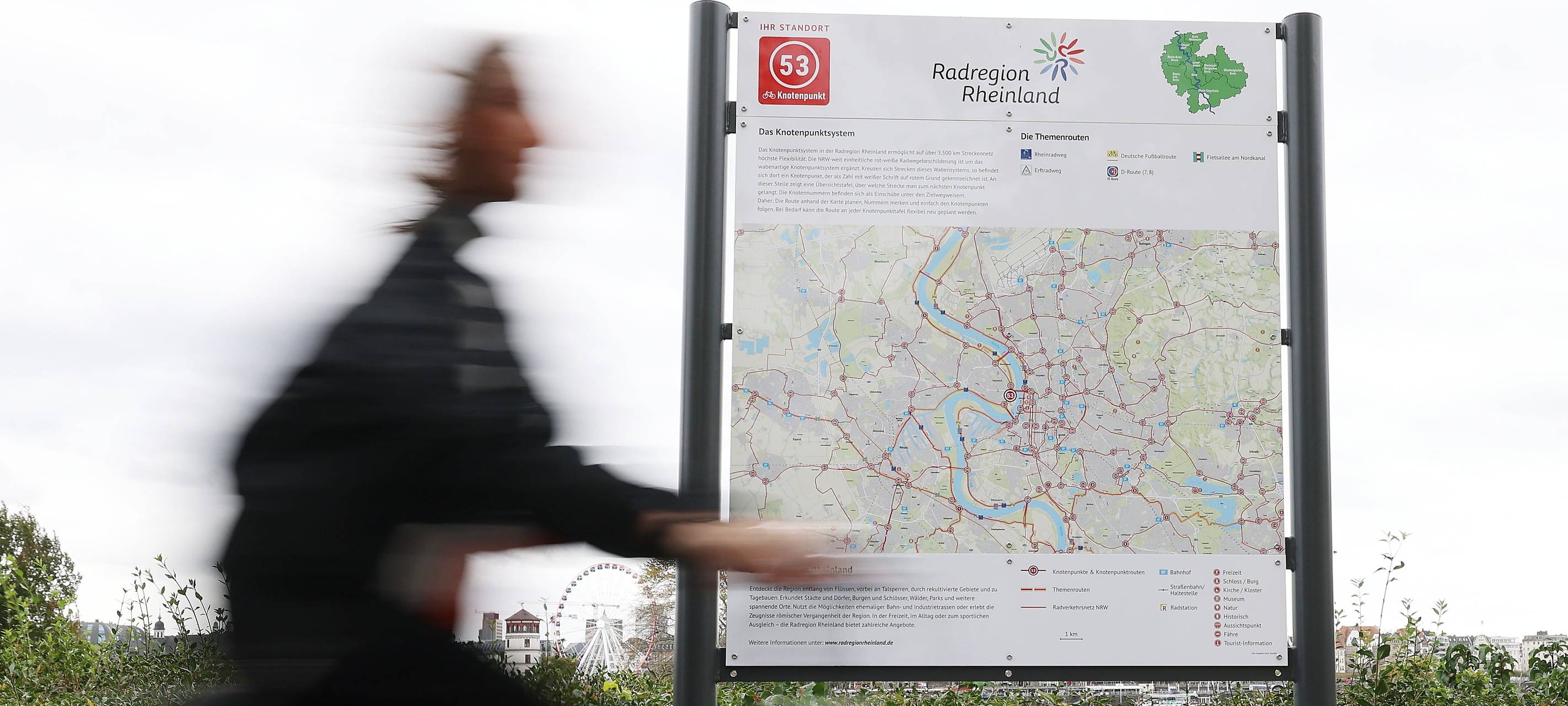 Vor einem Schild mit einer großen Fahrradkarte von Düsseldorf und der Region fährt ein Radfahrer entlang. Im Hintergrund sind der Schlossturm und das Riesenrad zu erkennen.