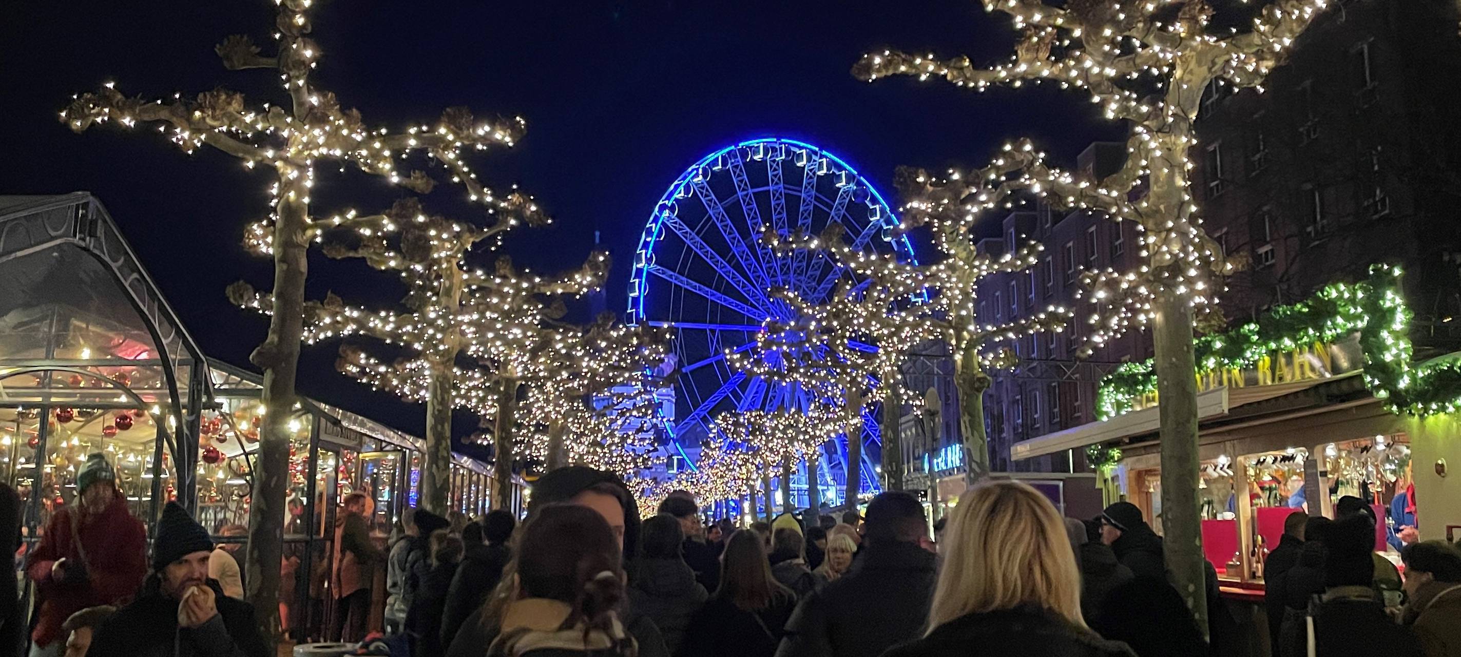 Lichterglanz auf dem Weihnachtsmarkt Düsseldorf am Abend". Menschen schlendern über den Roncalli-Weihnachtsmarkt in Düsseldorf. Im Hintergrund ist ein Riesenrad zu sehen.