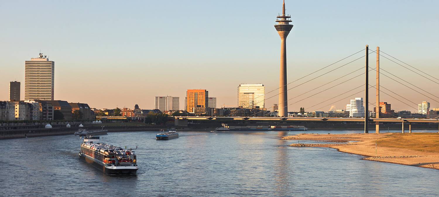 Links und im Vordergrund der Rhein, in dem ein Containerschiff schwimmt, rechts das Ufer. Im Hintergrund der Blick auf Düsseldorf, die Rheinkniebrücke und den Rheinturm.