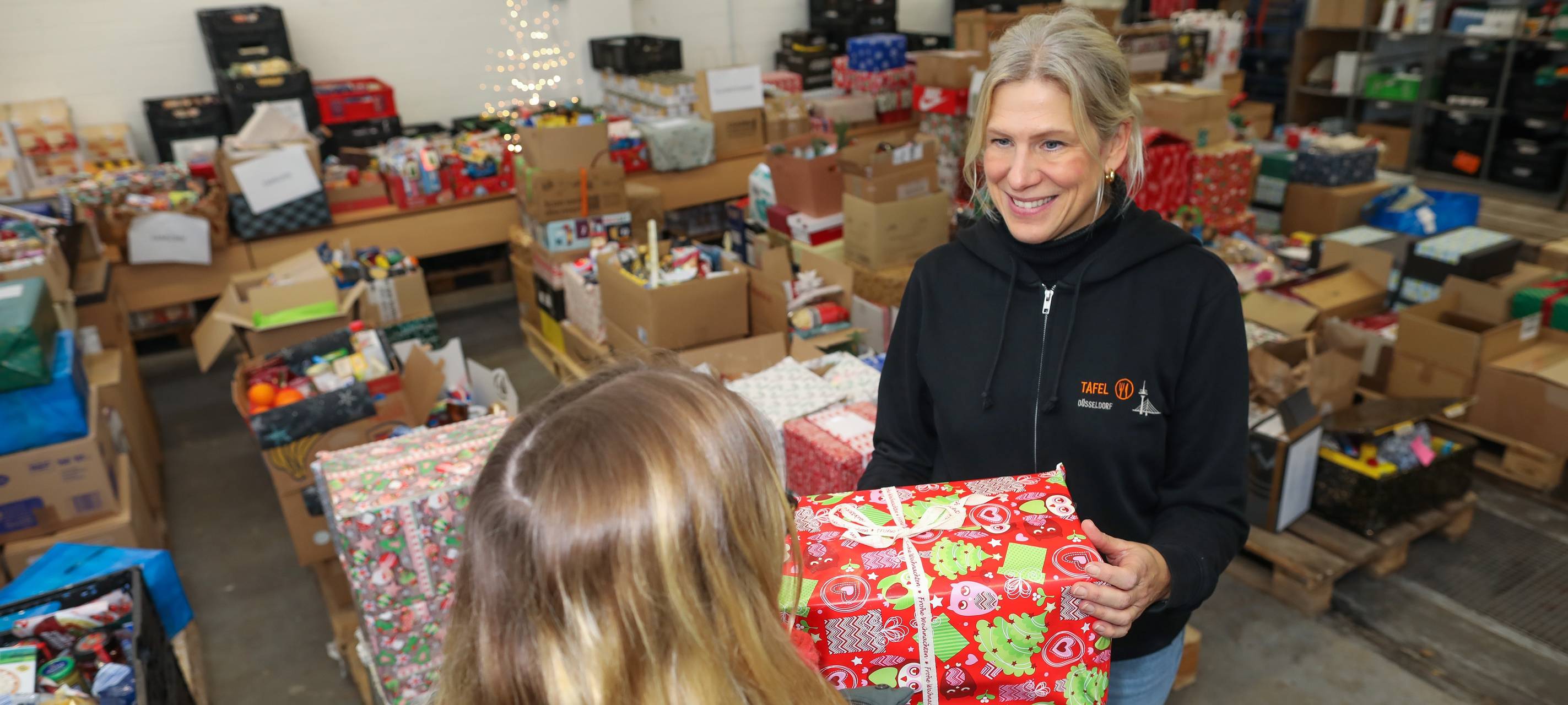 Tafel-Vorständin Eva Fischer übergibt ein Weihnachtspäckchen. In der Halle liegen viele weitere gespendete Päckchen.