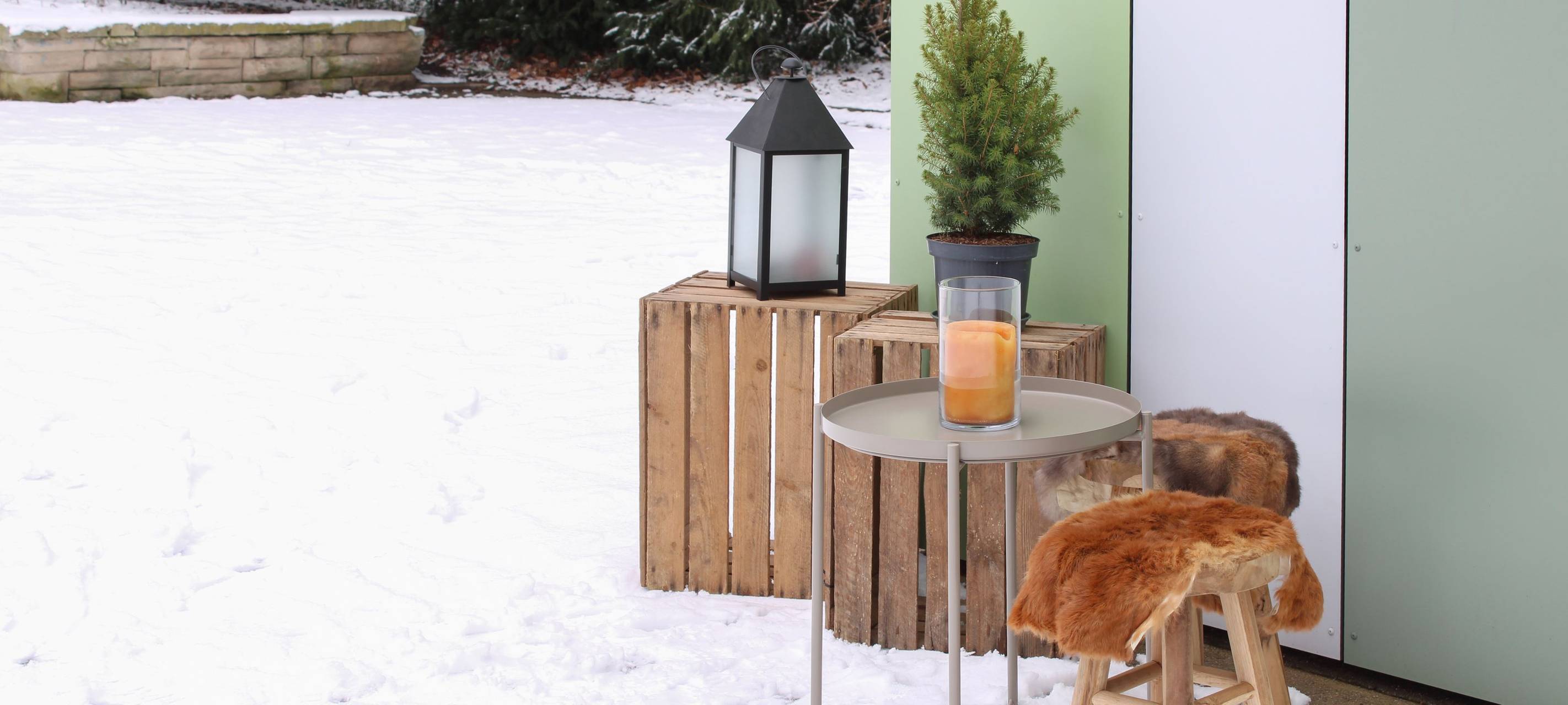 Ein kleiner Hocker mit Fell, ein Beistelltisch und zwei Kisten mit einer Laterne darauf vor der Ecke einer grünen Hütte im zugeschneiten Sternwartpark.