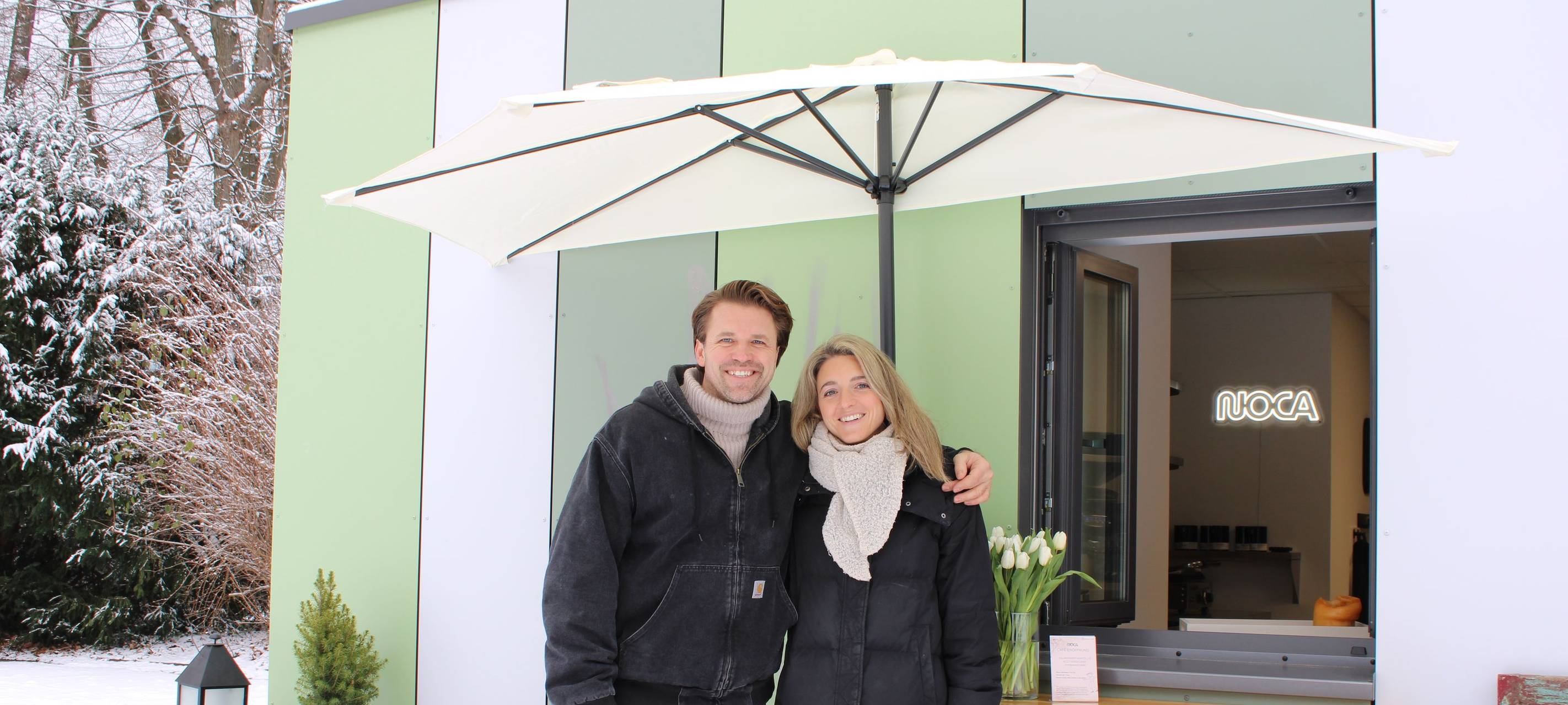 Ein Mann und eine Frau stehen unter einem Schirm vor einer grünen Hütte in einem Park.
