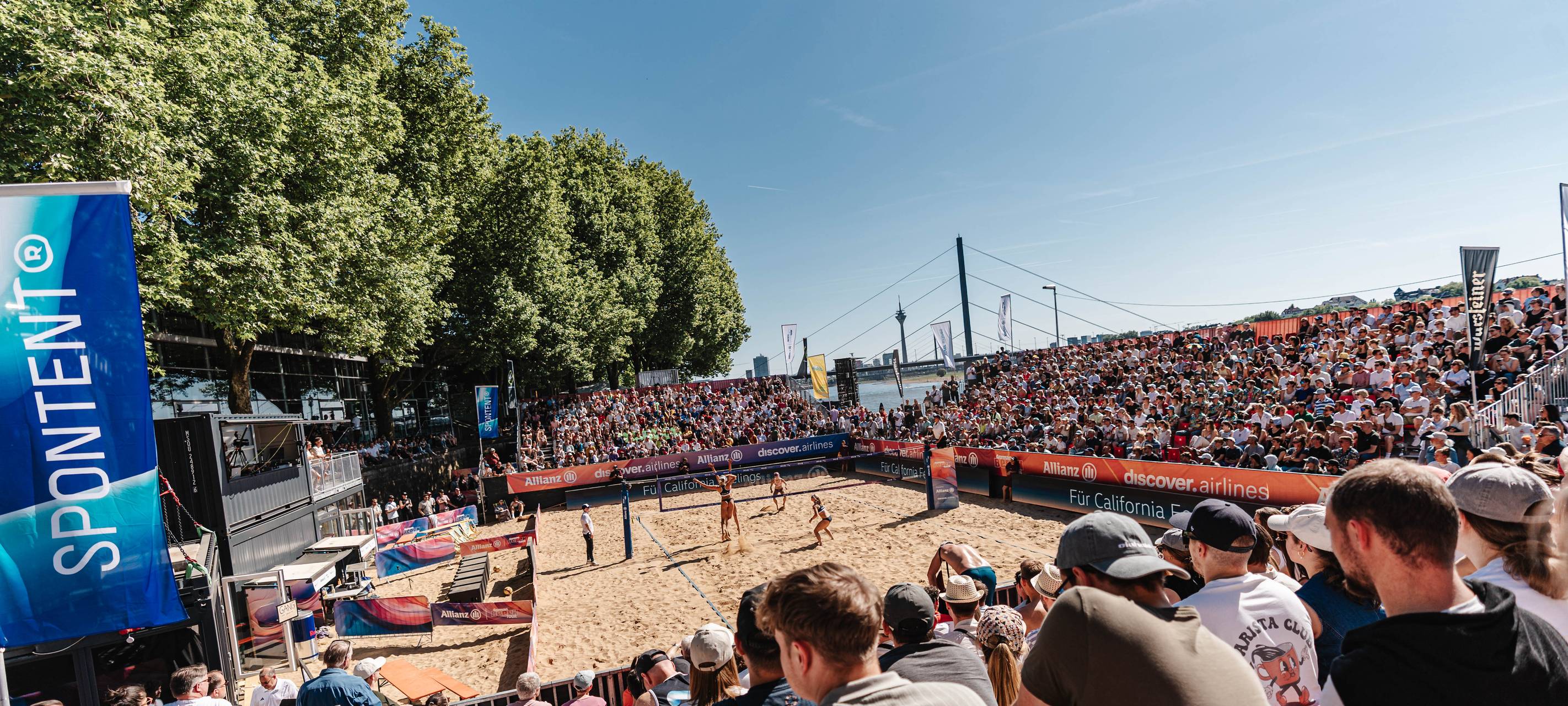 Beachvolleyballarena in Düsseldorf mit Publikum. Im Hintergrund sind der Rhein, die Kniebrücke und der Rheinturm zu sehen.