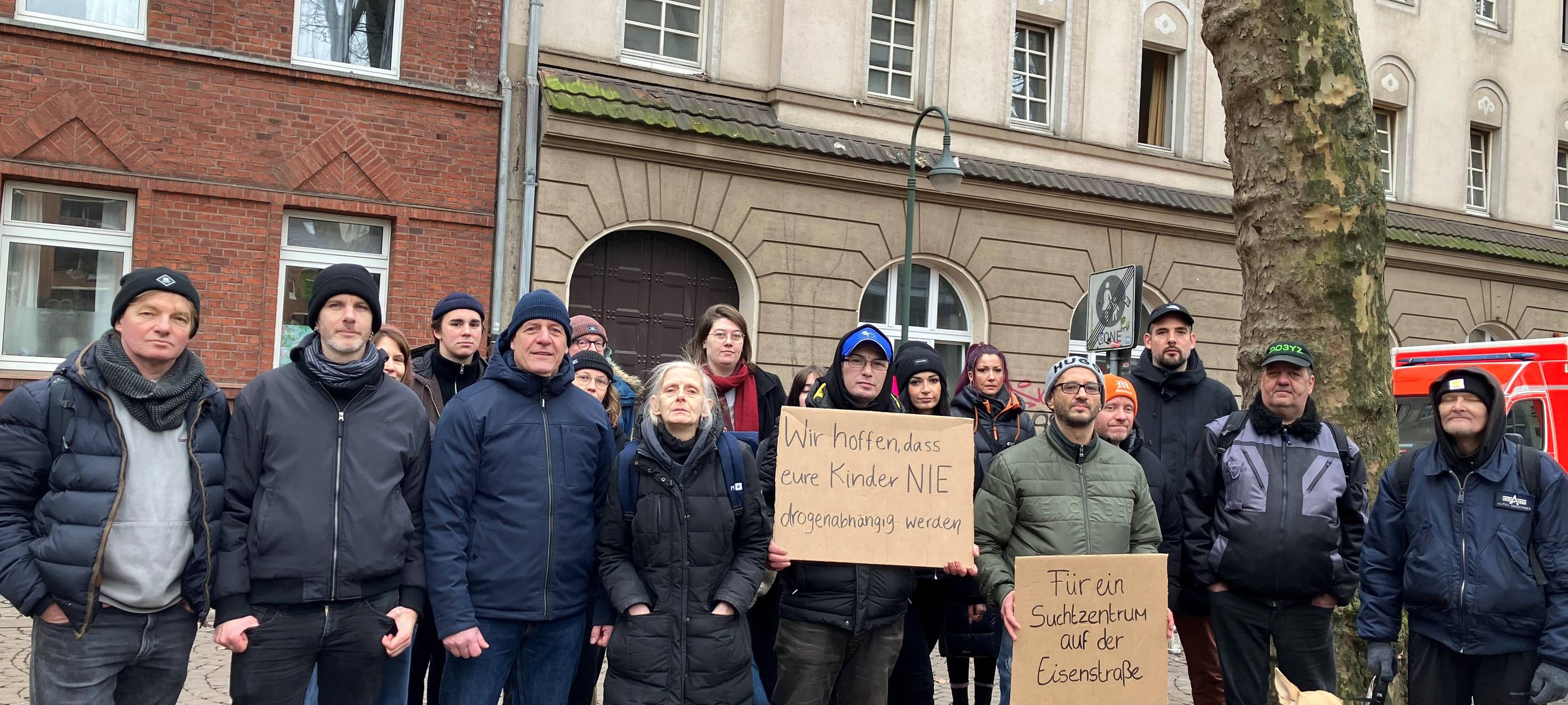 Eine Gruppe Menschen steht vor dem Gebäude "Eisenstraße 49". Einige halten Schilder in der Hand. Darauf steht "Für ein Suchtzentrum auf der Eisenstraße".