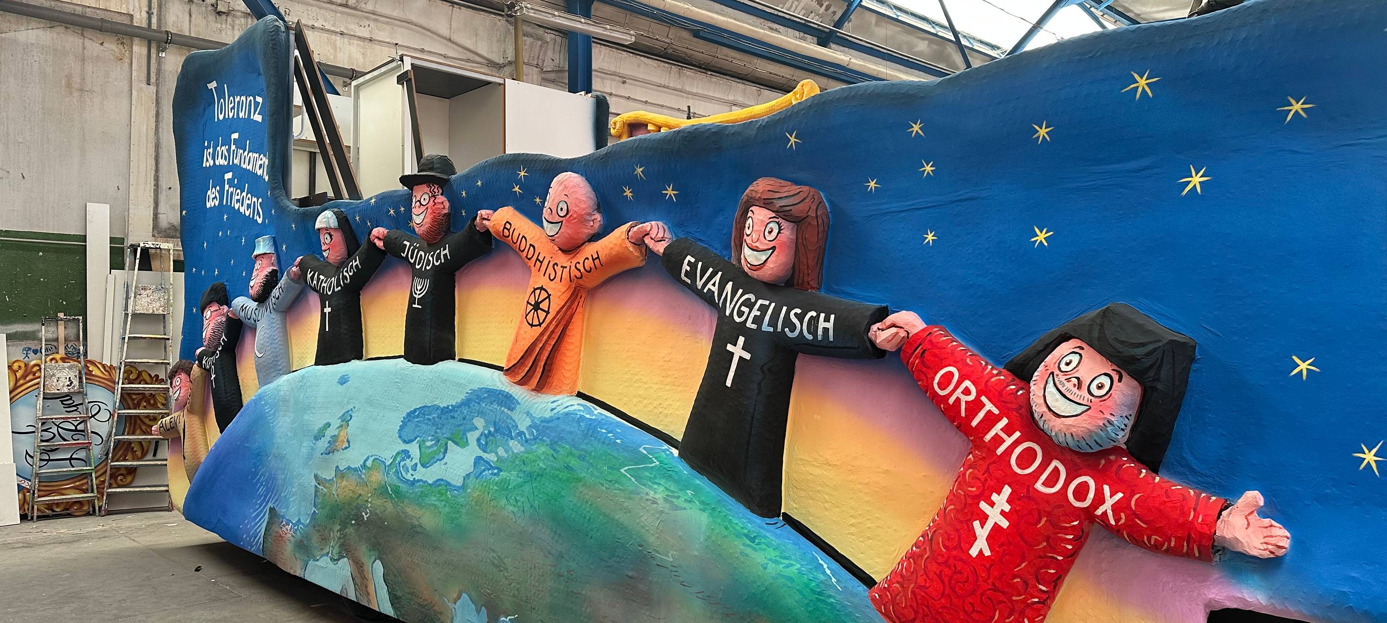 Der Toleranz-Karnevalswagen steht in der Wagenbauhalle. Verschiedene Personen aus verschiedenen Religionen halten die Hand.