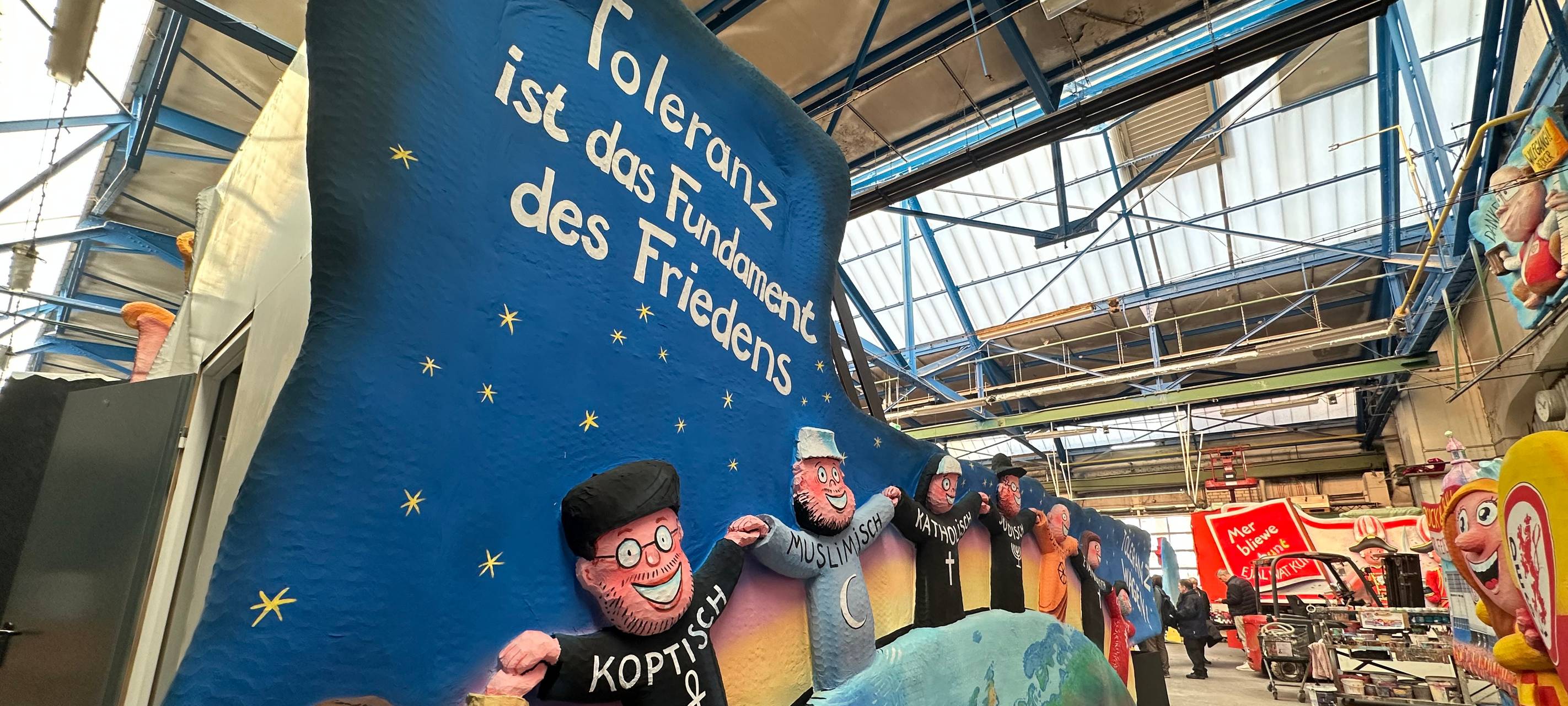 Der Toleranz-Karnevalswagen steht in der Wagenbauhalle. Verschiedene Personen aus verschiedenen Religionen halten die Hand.