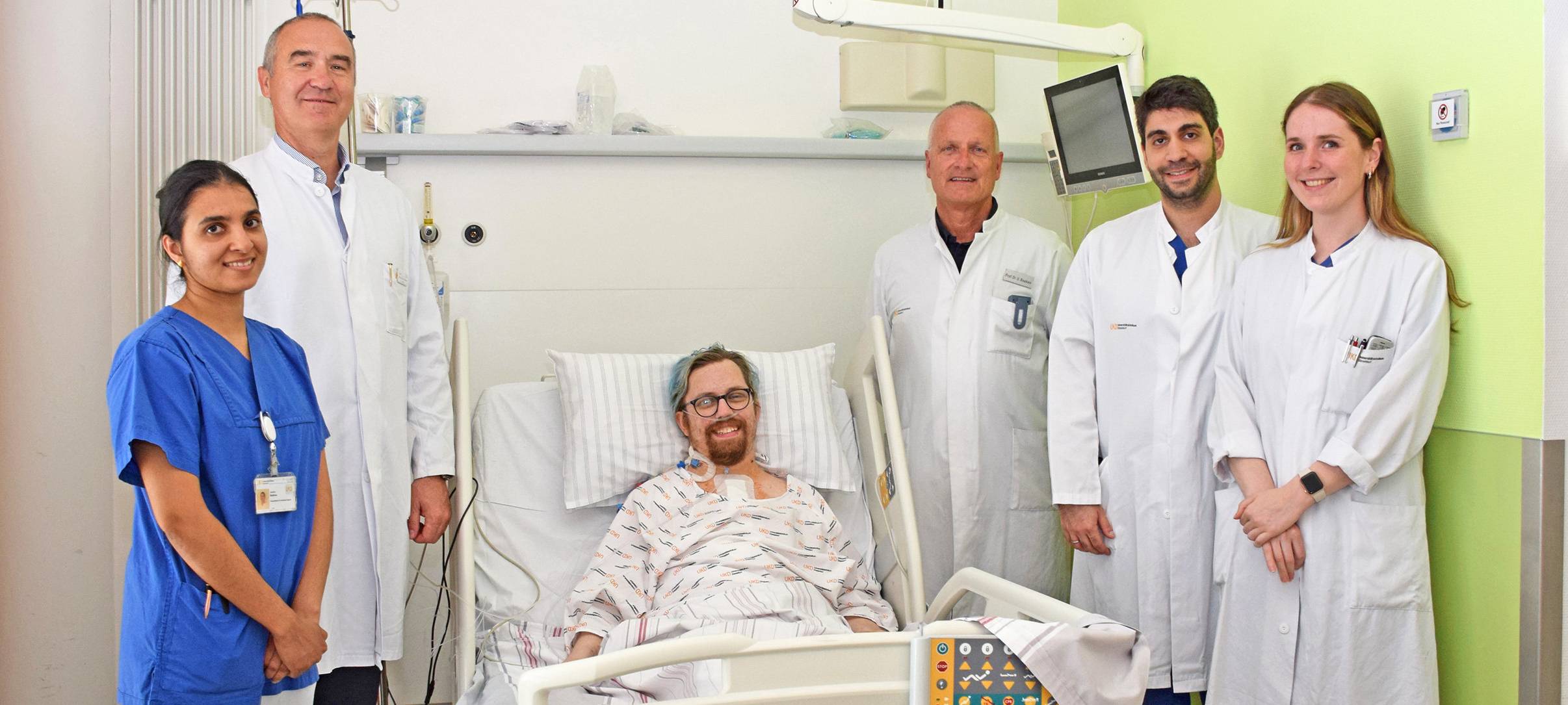 5 Menschen in weißen Kitteln stehen um ein Krankenbett. Darin liegt ein Patient. Alle lächeln in die Kamera.