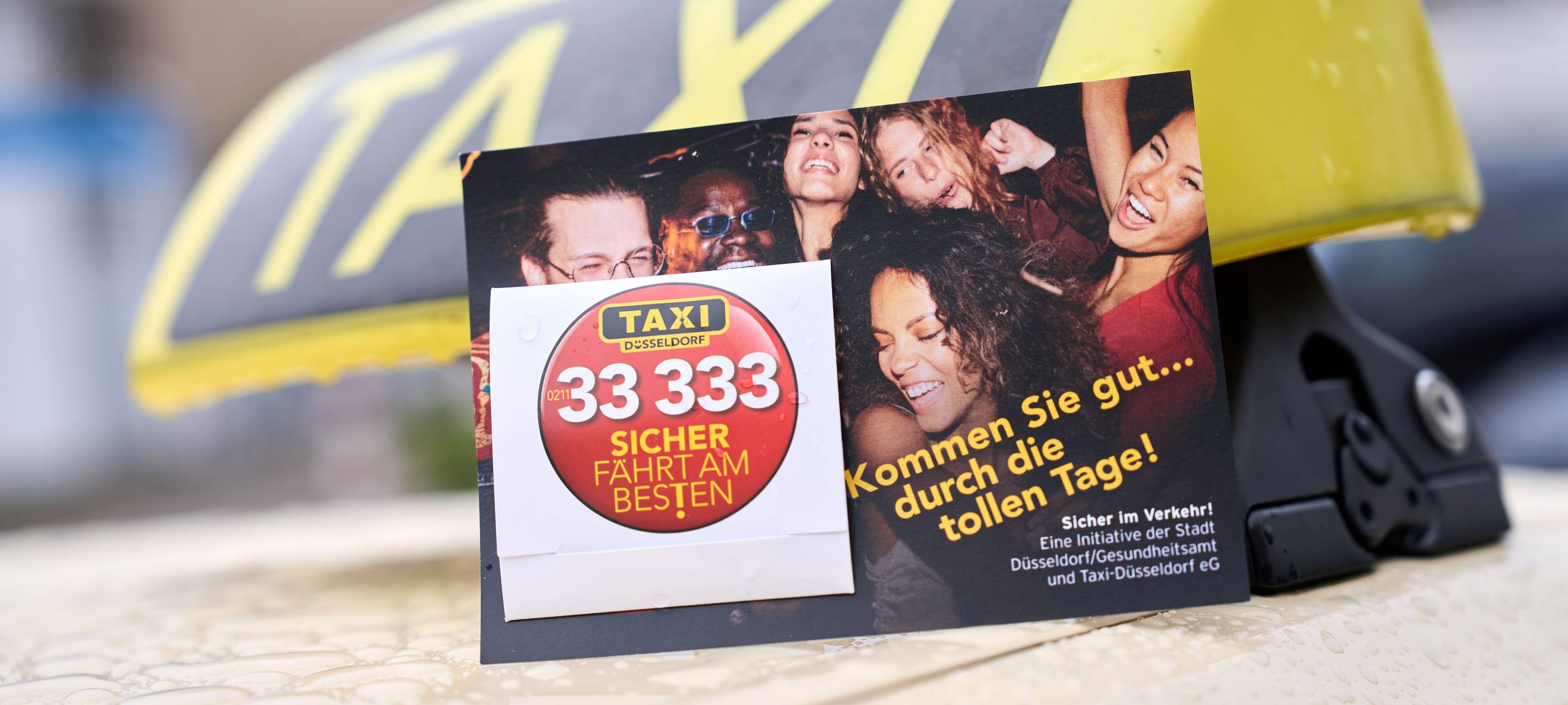 Nahaufnahme eines Taxidaches. An dem Taxischild lehnen Postkarten, an denen jeweils ein Kondom befestigt ist.