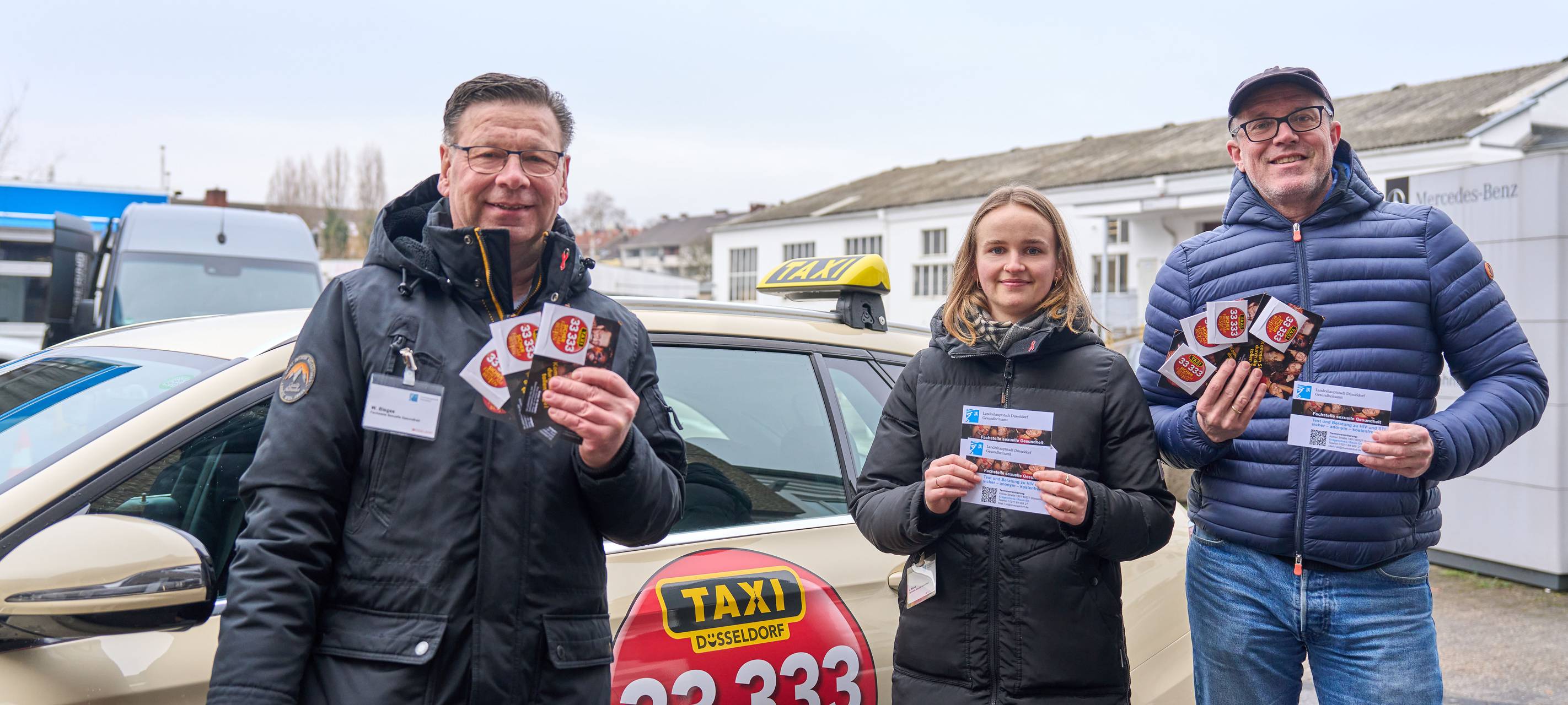 Zwei Männer und eine Frau stehen vor einem Taxi. Sie halten eine Broschüre bzw. eine Postkarte mit je einem Kondom in der Hand und lächeln in die Kamera.