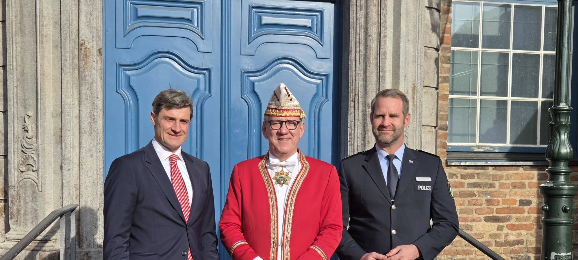 Drei Männer stehen vor der blauen Tür des Rathauses. In der Mitte ein Mann in Karnevalsuniform, die Männer außen tragen Anzüge.