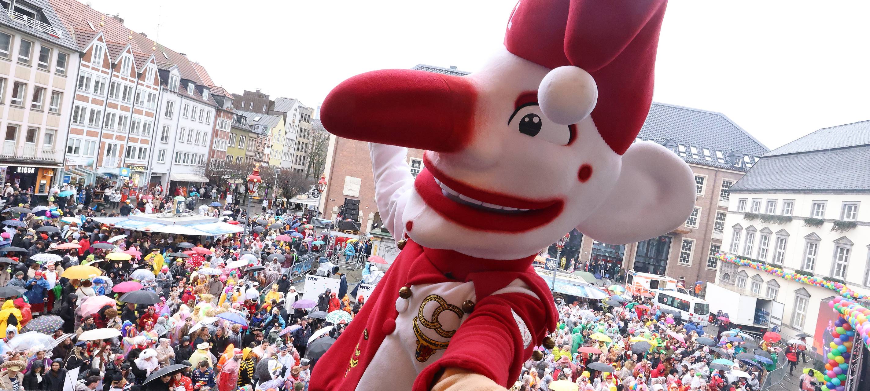Die Figur des Hoppeditz steht an Altweiber auf dem Rathaus-Balkon. Vor dem Rathaus feiern währenddessen viele Jecke trotz Regenwetter.