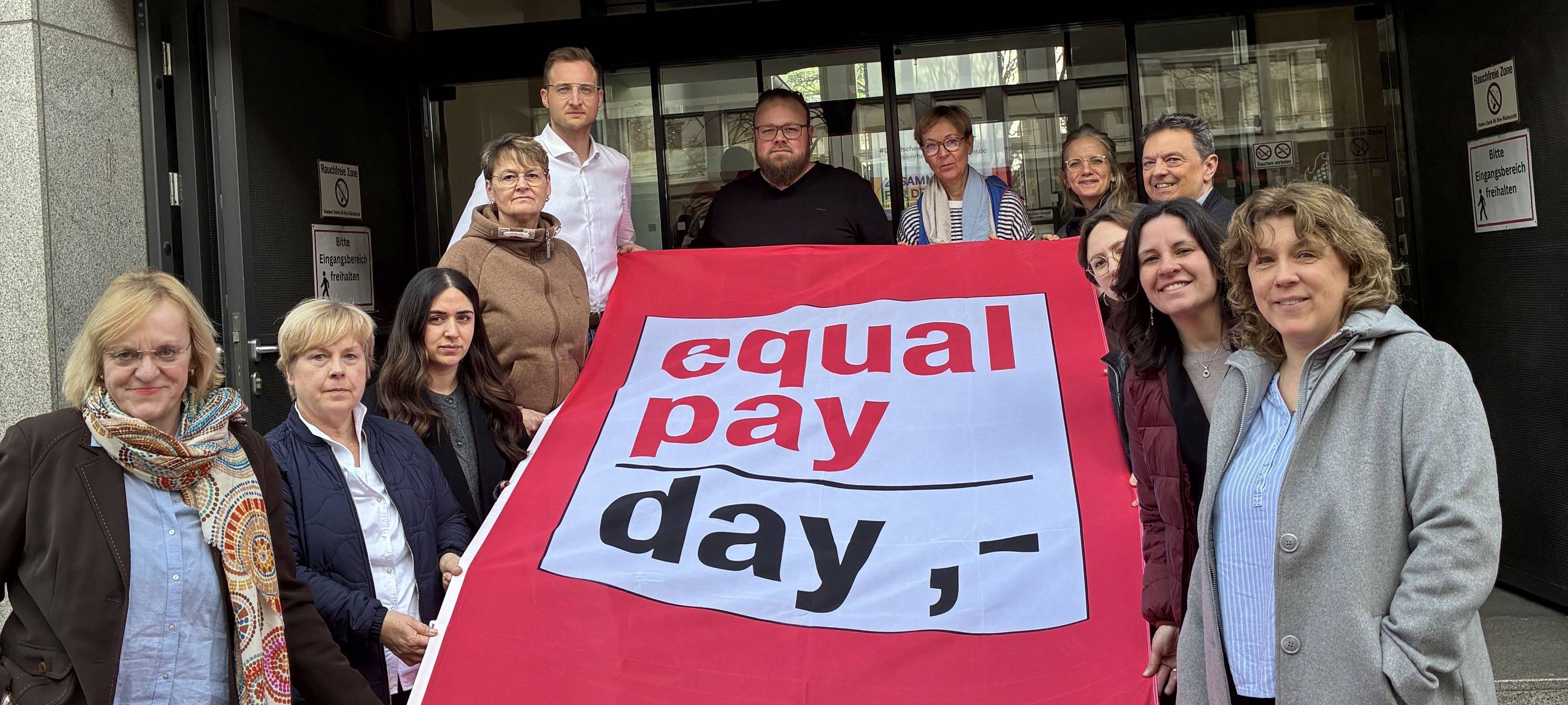 Mitglieder des DGB stehen mit einer roten Flagge vor ihrem Eingang und weisen auf den Equal Pay Day hin