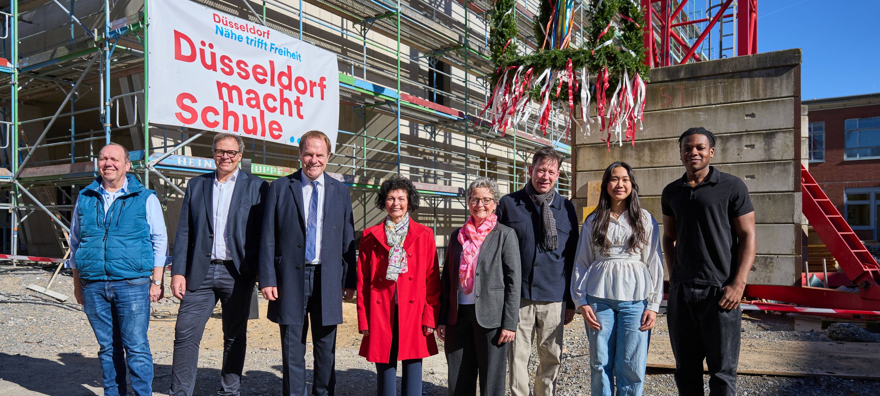 OB Keller und andere verantwortliche Frauen und Männer stehen vor dem Rohbau der Joseph-Beuys-Gesamtschule. Hinter ihnen schwebt der Kranz für das Richtfest.