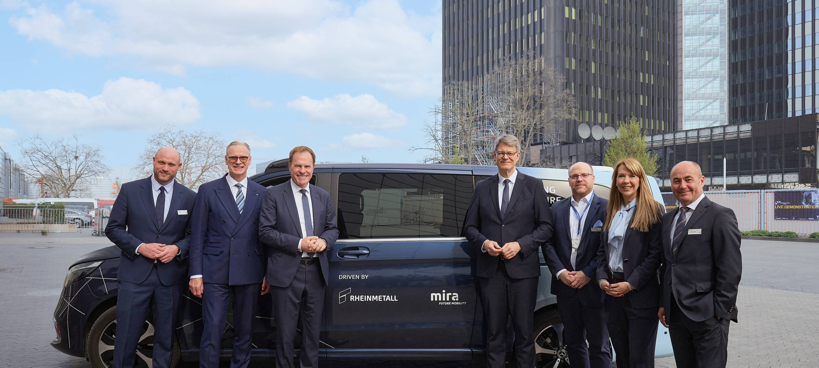 Sechs Männer und eine Frau in Business-Kleidung stehen vor einem schwarzen Fahrzeug. Darauf sind die Logos von "Rheinmetall" und "MIRA" zu sehen.