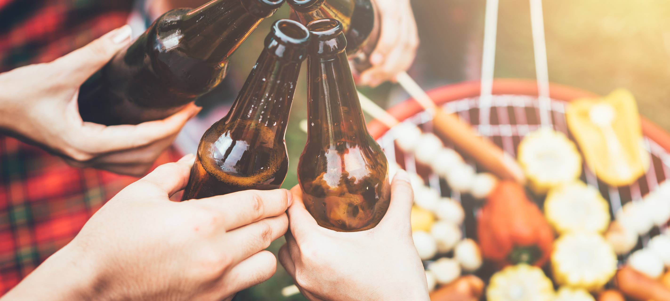 Nahaufnahme von vier Personen, die mit Bierflaschen über einem brennenden Grill anstoßen. Auf dem Grill liegen Maiskolben, Paprika und Würstchen. Warme, sonnige Atmosphäre im Freien.