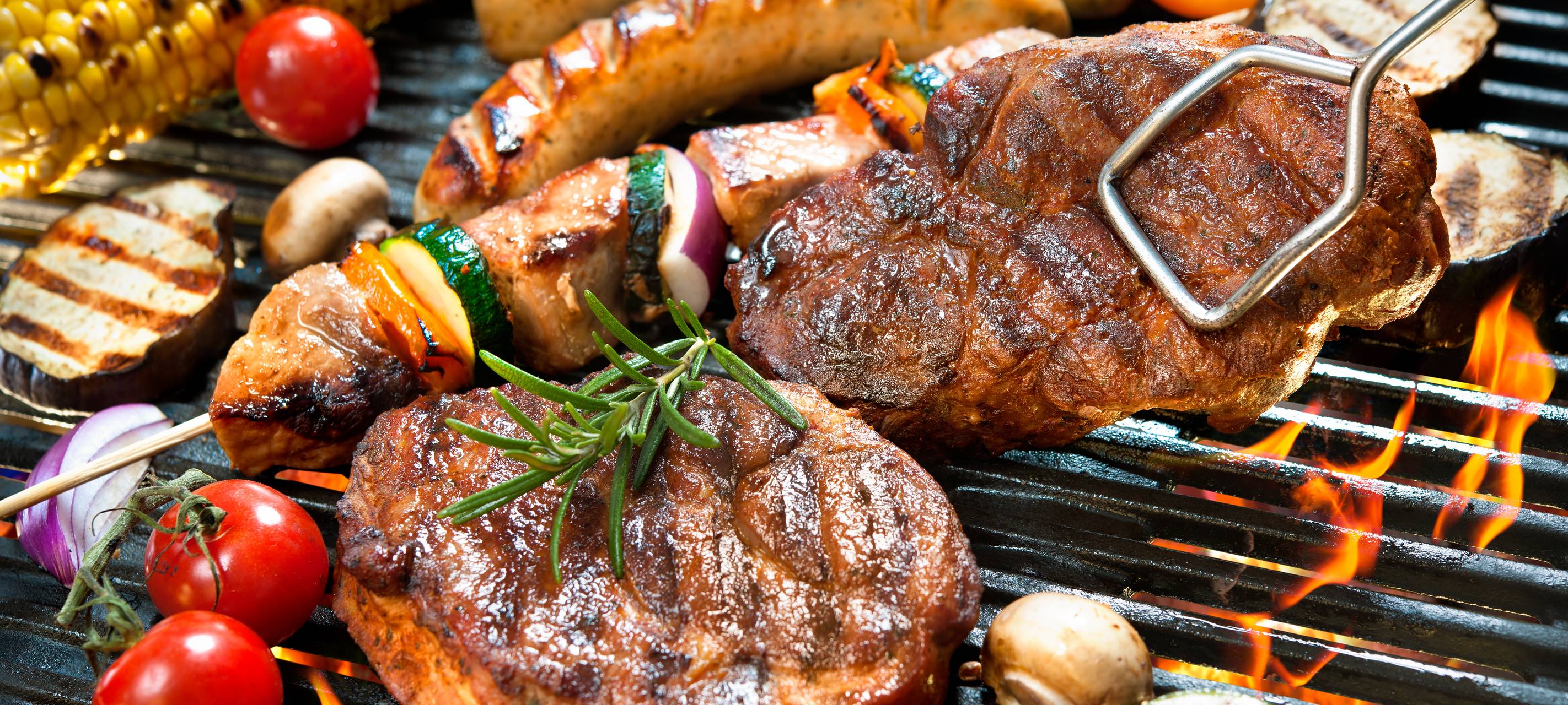 Nahaufnahme von verschiedenen Fleischsorten wie Steaks und Würstchen sowie buntem Grillgemüse (Mais, Tomaten, Spieße) auf einem Grillrost über offenen Flammen.