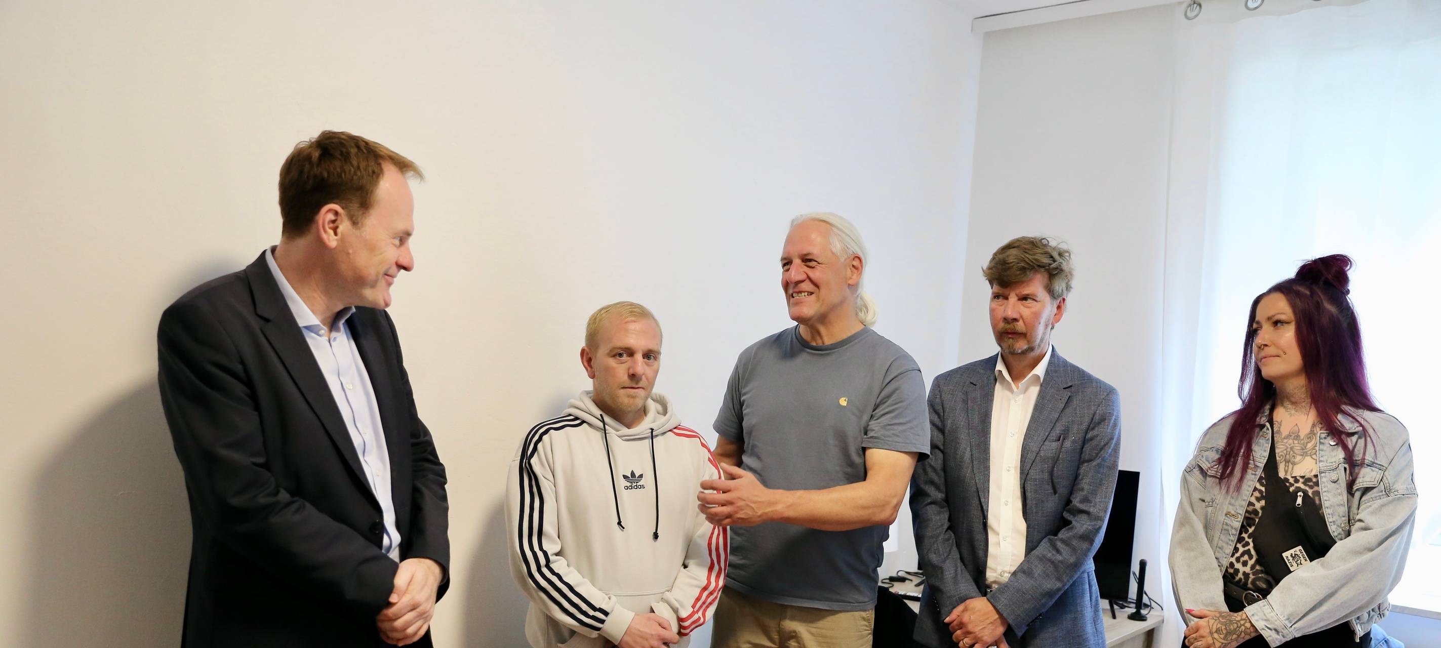 Oberbürgermeister Stephan Keller, Wohnungsnehmer Thomas D., Oliver Ongaro von FiftyFifty und weitere Personen stehen vor einer weißen Wand in Thomas' neuer Wohnung