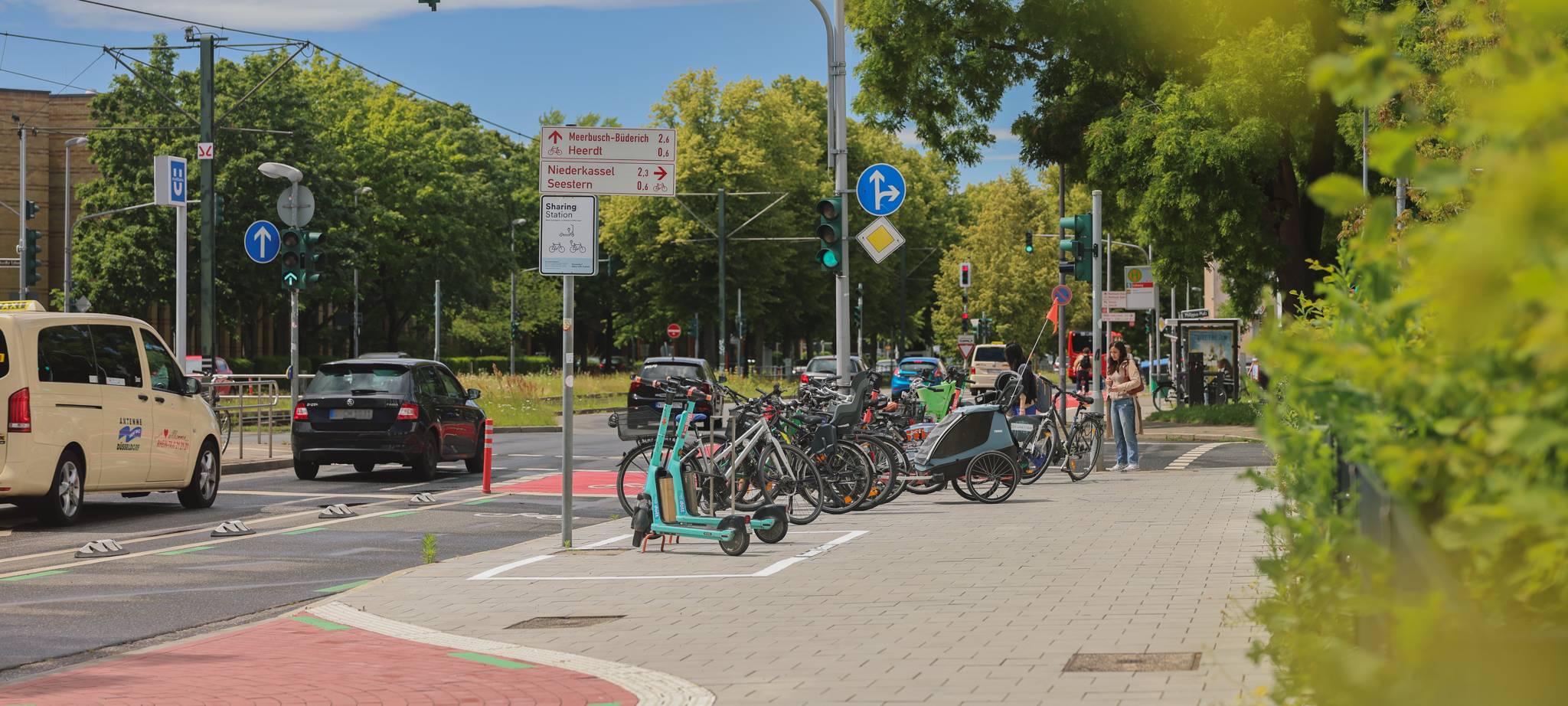 E-Scooter stehen in einem markierten Bereich auf einem breiten Gehweg, daneben das Schild "Sharing-Station". Dahinter stehen zahlreiche Fahrräder in Fahrradständern.