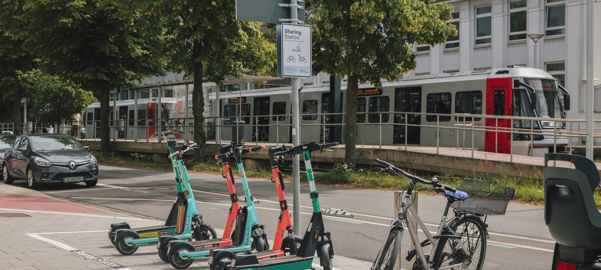 Auf dem Gehweg ist eine markierte Sharingstation, an der mehrere E-Scooter geparkt sind. Im Hintergrund hält gerade eine Niederflurbahn am Bahnsteig.