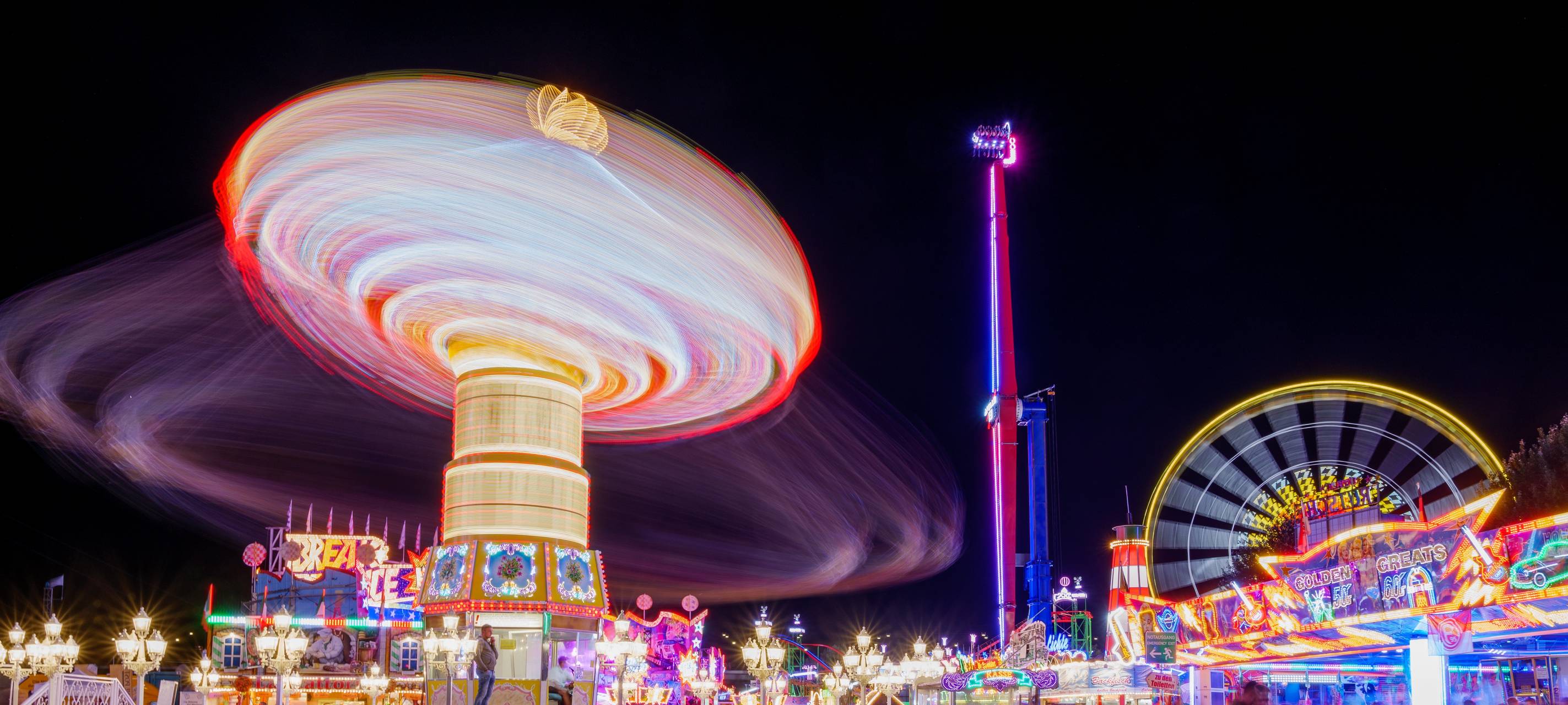Nächtliche Stimmung auf der Rheinkirmes mit farbig beleuchtetem Karussell in Bewegung und Riesenrad im Hintergrund. So oder ähnlich wird es auch bei der Rheinkirmes 2026 aussehen.