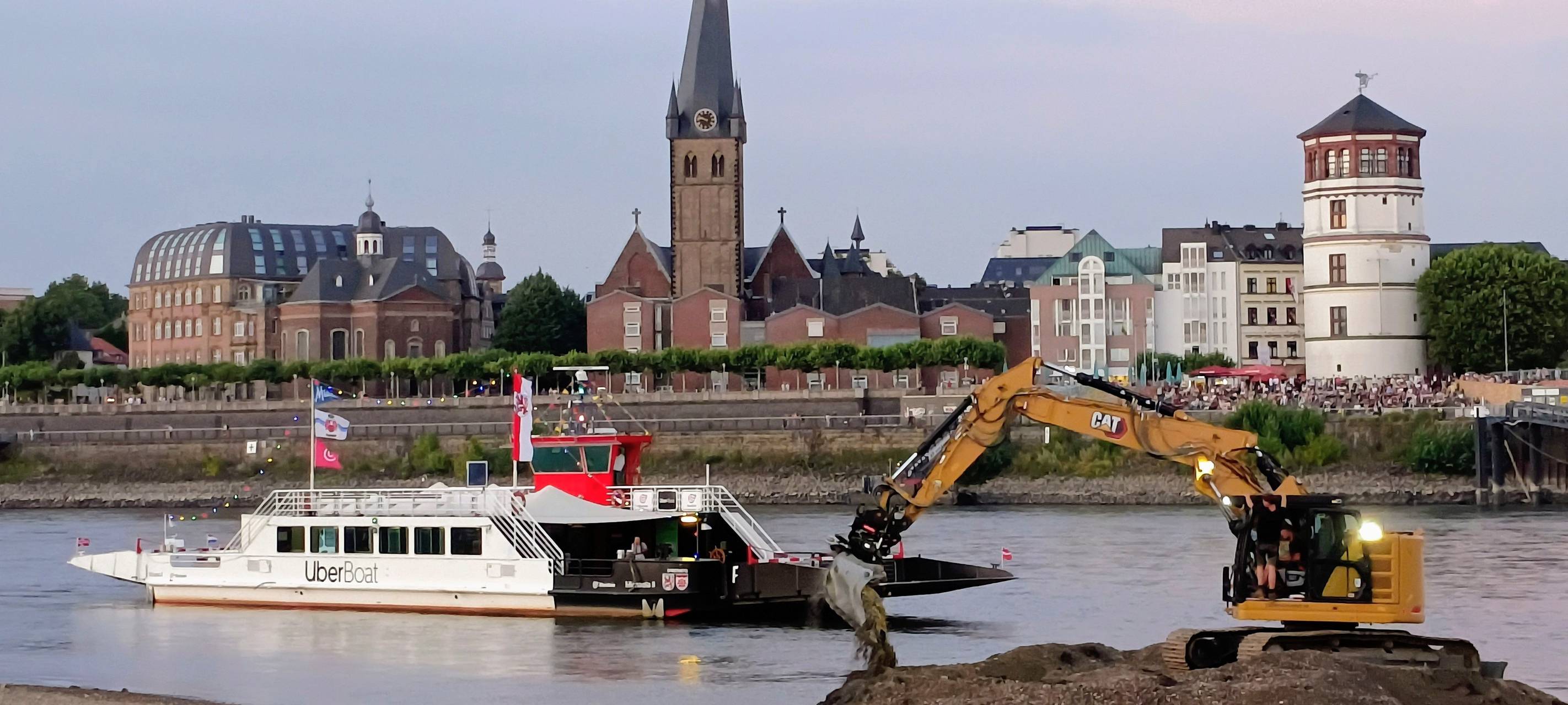 Ein Bagger im Einsatz am Rhein. Im Hintergrund die Fähre, die Menschen zur Rheinkirmes transportieren soll. Am ersten Wochenende der Kirmes konnte die Fähre wegen des Niedrigwasser nicht fahren.