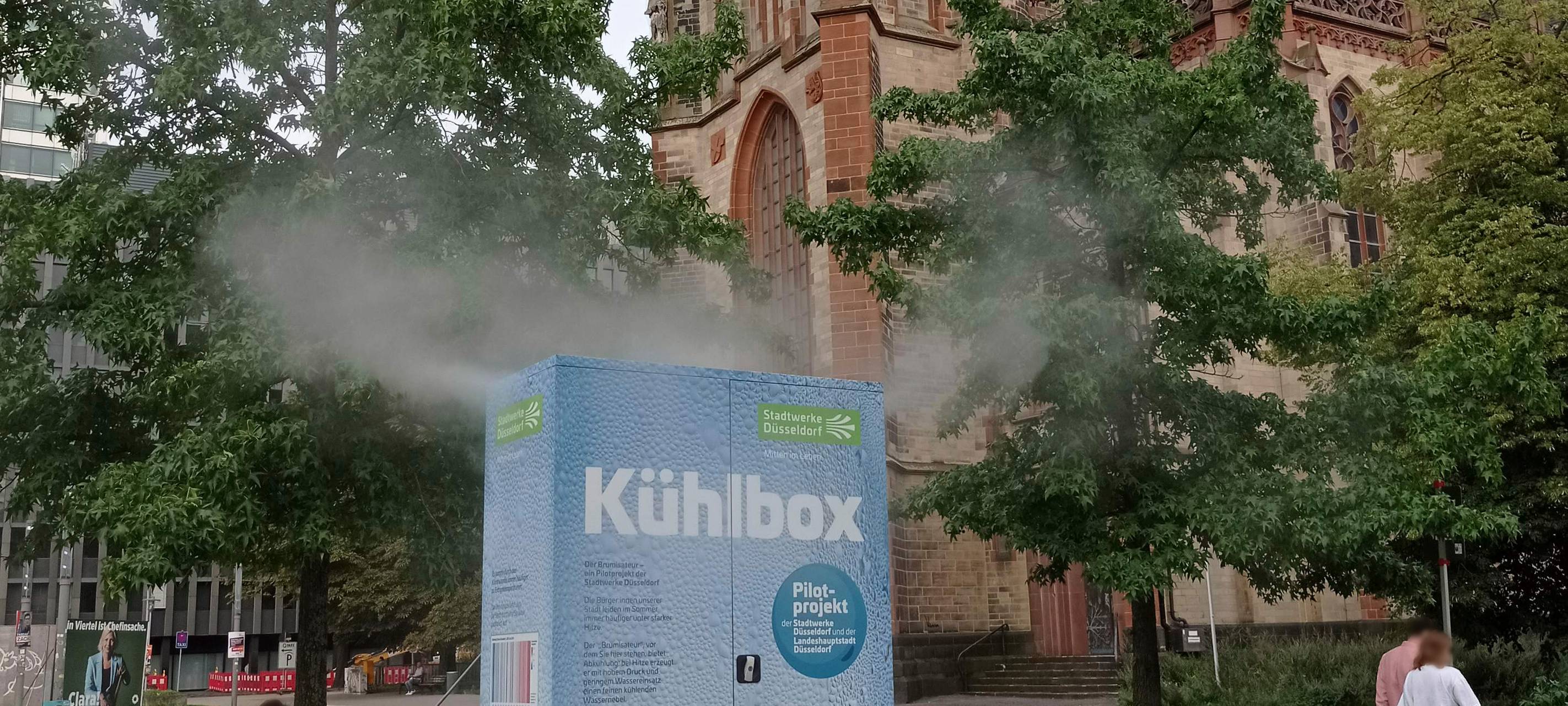Die Kühlbox "Brumisateur" auf dem Kirchplatz versprüht Wassernebel. Im Hintergrund ist die Kirche St. Peter zu sehen.