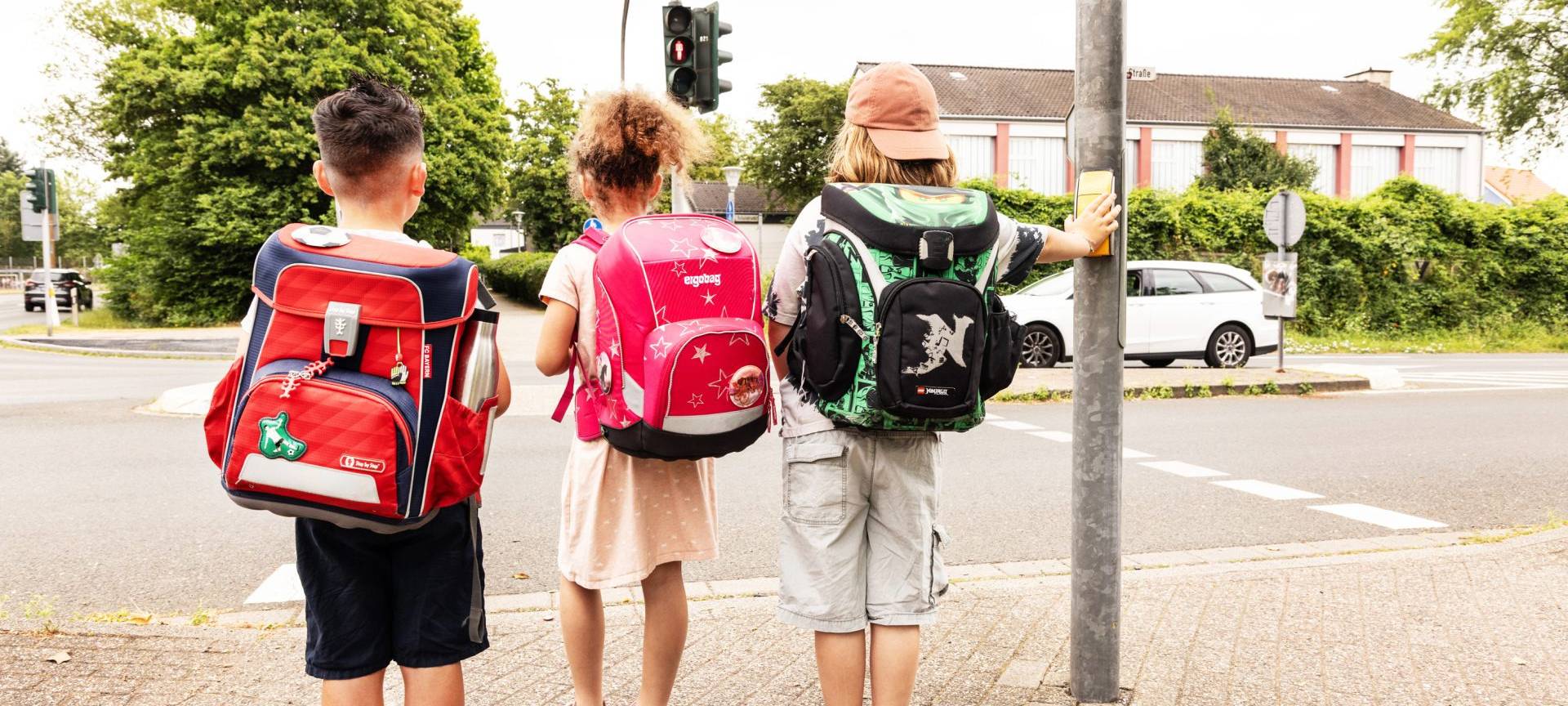 Drei Schulkinder stehen auf ihrem Schulweg an einer roten Ampel.