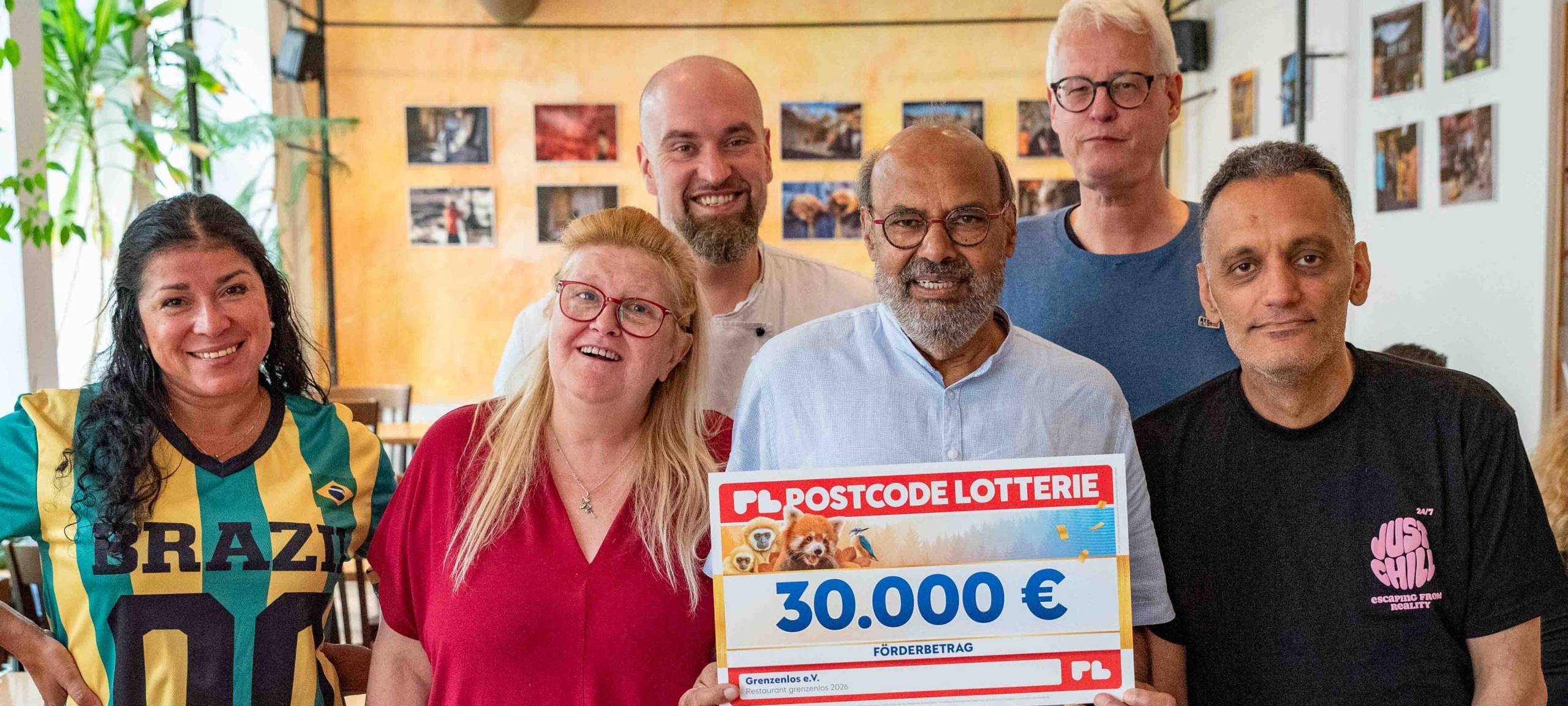 Sechs Menschen stehen in einem Raum und halten einen Scheck der Deutschen Postcode Lotterie in die Kamera.
