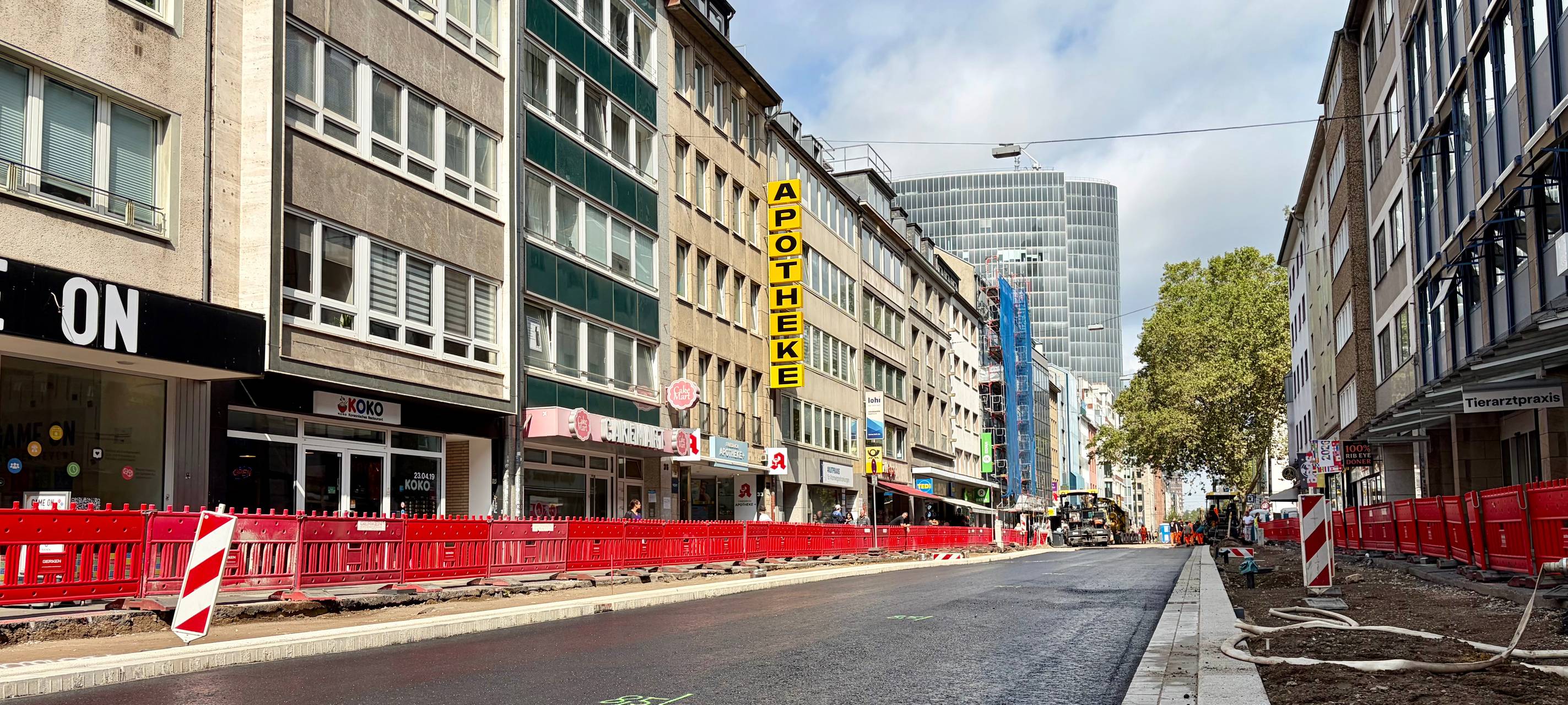 Die frisch asphaltierte Friedrichstraße zwischen der Herzogstraße und dem Graf-Adolf-Platz. Die Straße ist noch mit Baken begrenzt. Auf der linken Seite befinden sich eine Apotheke und andere Geschäfte.