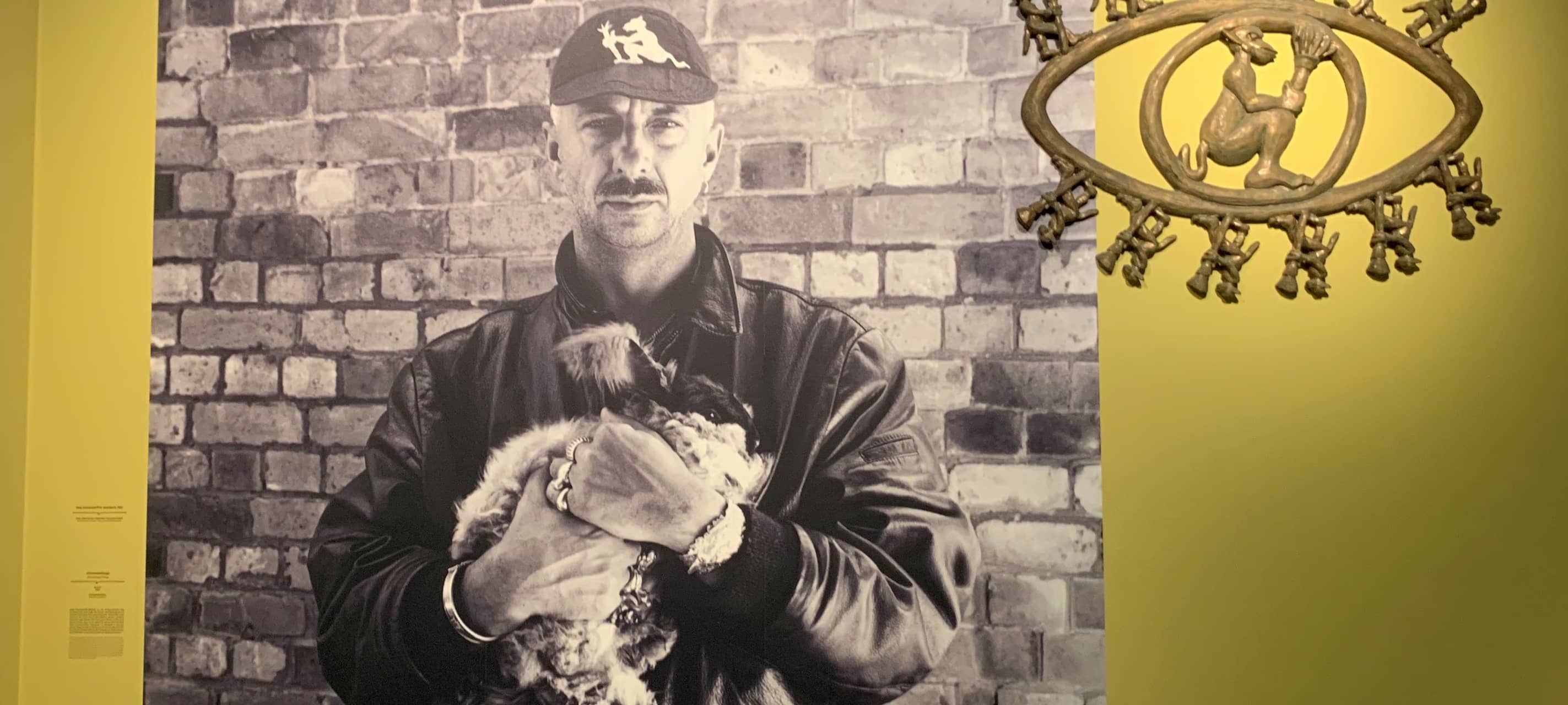Aufnahme von einem Bild. Auf dem Bild steht ein Mann vor einer Steinmauer. Er trägt eine schwarze Lederjacke und hällt einen Hund in der Hand.