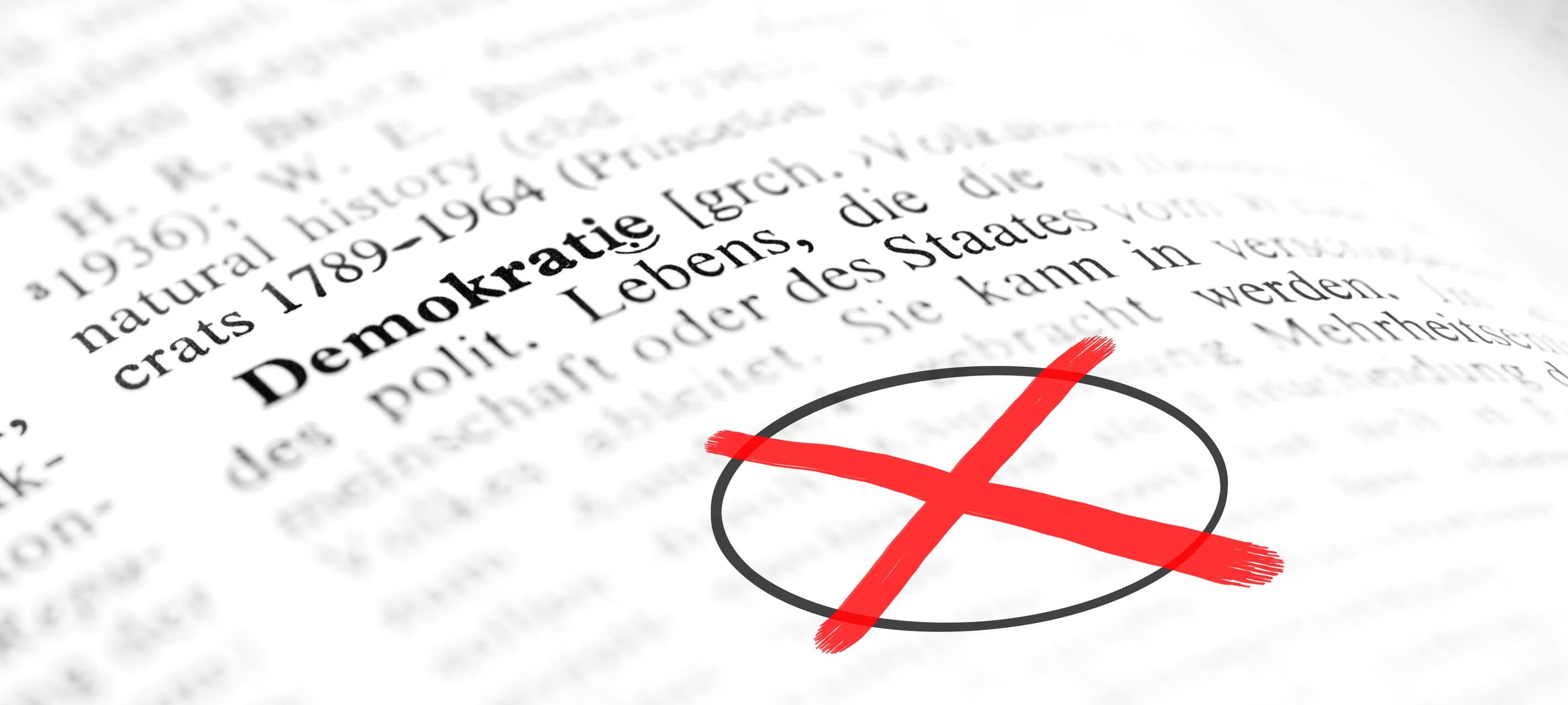 Eine Nahaufnahme einer gedruckten Textseite: Das Wort „Demokratie“ steht im Vordergrund, während der übrige Text unscharf bleibt. Daneben ist ein Bereich mit einem schwarzen Kreis markiert und mit einem auffälligen roten Kreuz durchgestrichen.