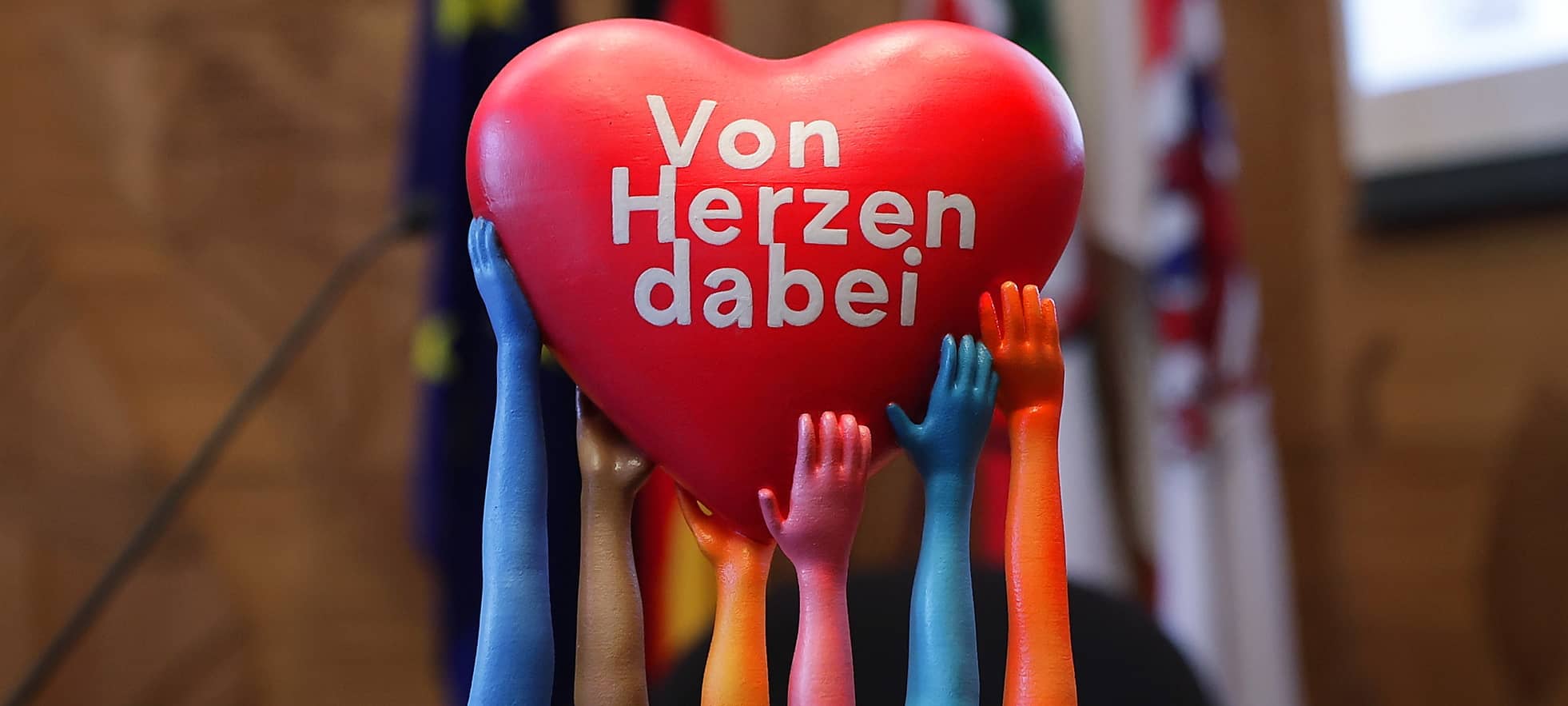 Auf einem roten Herz steht in weißer Schrift: "Von Herzen dabei". Getragen wird das Herz von mehreren Armen in verschiedenen Farben.