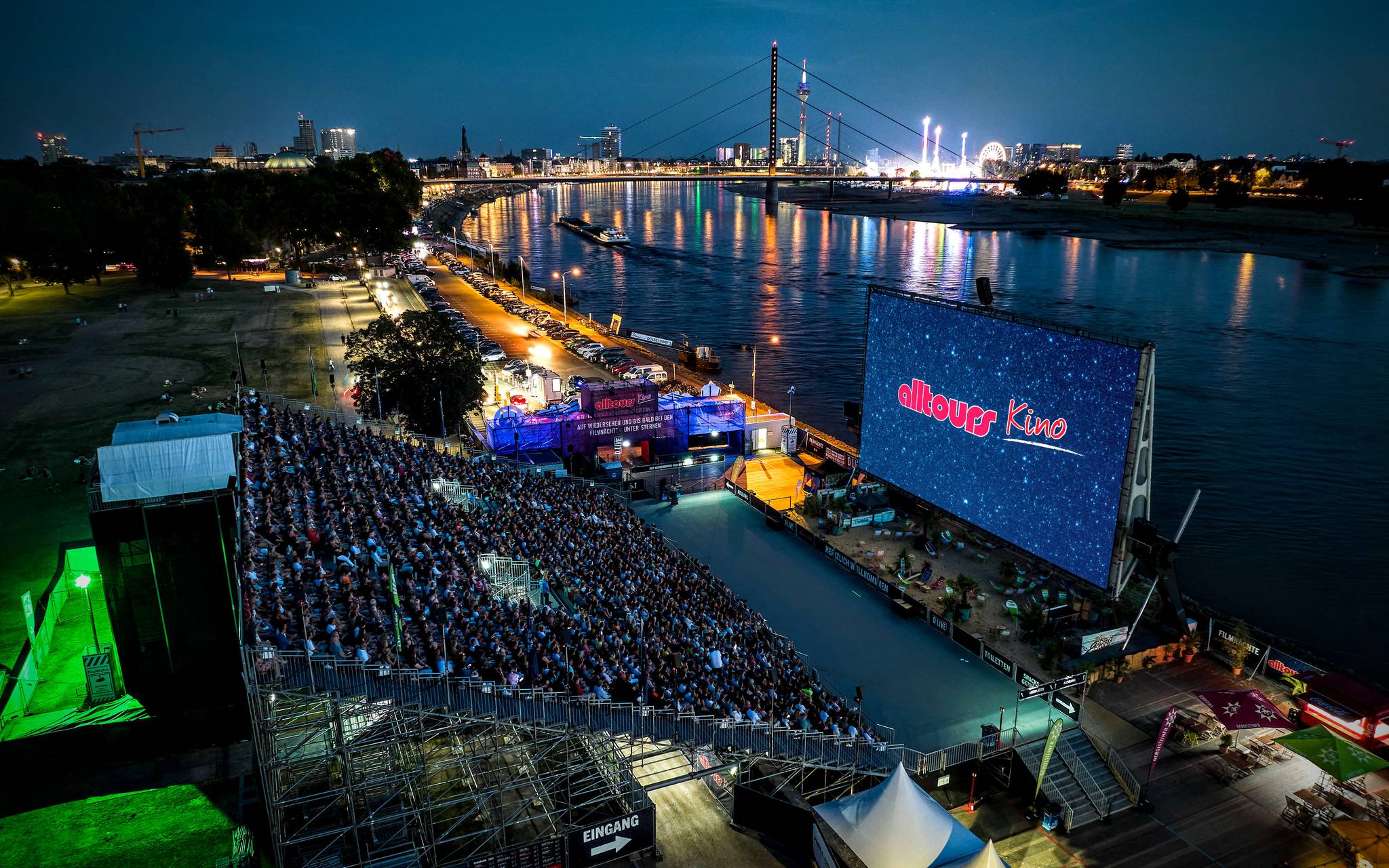 Zu sehen ist das voll besetzte alltours Open Air Kino direkt am Rhein, auf der Leinwand ist das alltours Logo zu sehen. Im Hintergrund kann man die Skyline Düsseldorfs mit dem Fernsehturm, der Rheinkniebrücke und der Rheinkirmes erkennen.