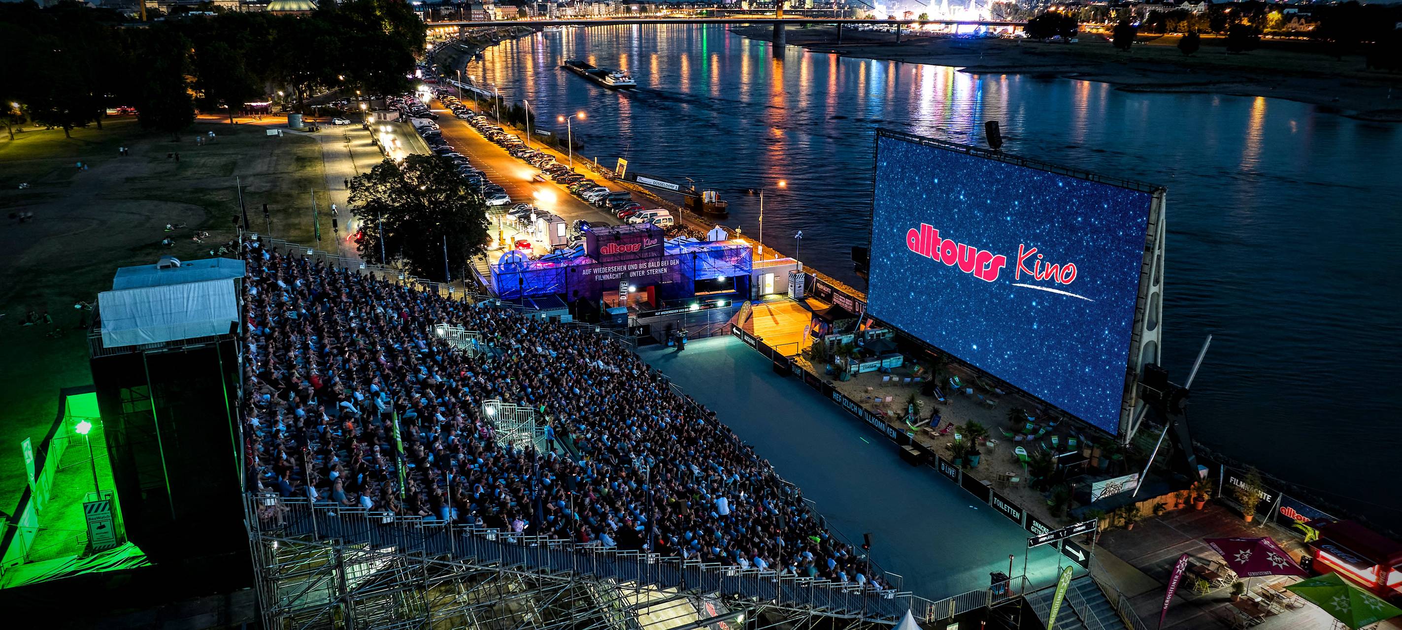 Zu sehen ist das voll besetzte alltours Open Air Kino direkt am Rhein, auf der Leinwand ist das alltours Logo zu sehen. Im Hintergrund kann man die Skyline Düsseldorfs mit dem Fernsehturm, der Rheinkniebrücke und der Rheinkirmes erkennen.