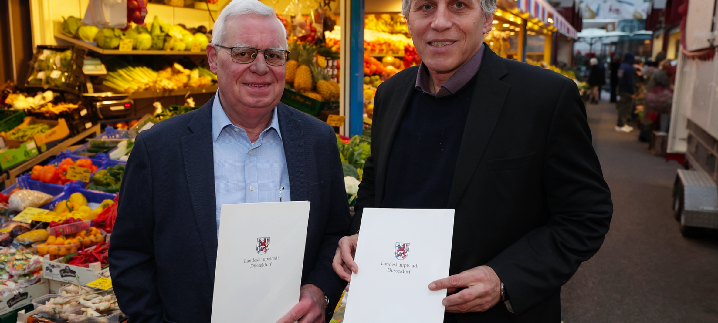 Umweltdezernent Jochen Kral und Carlsplatz-Chef Heiner Röckrath stehen vor einem Marktstand des Carlsplatzes. Sie präsentieren den neuen Mietvertrag, der die Zukunft des Carlsplatzes sichert.